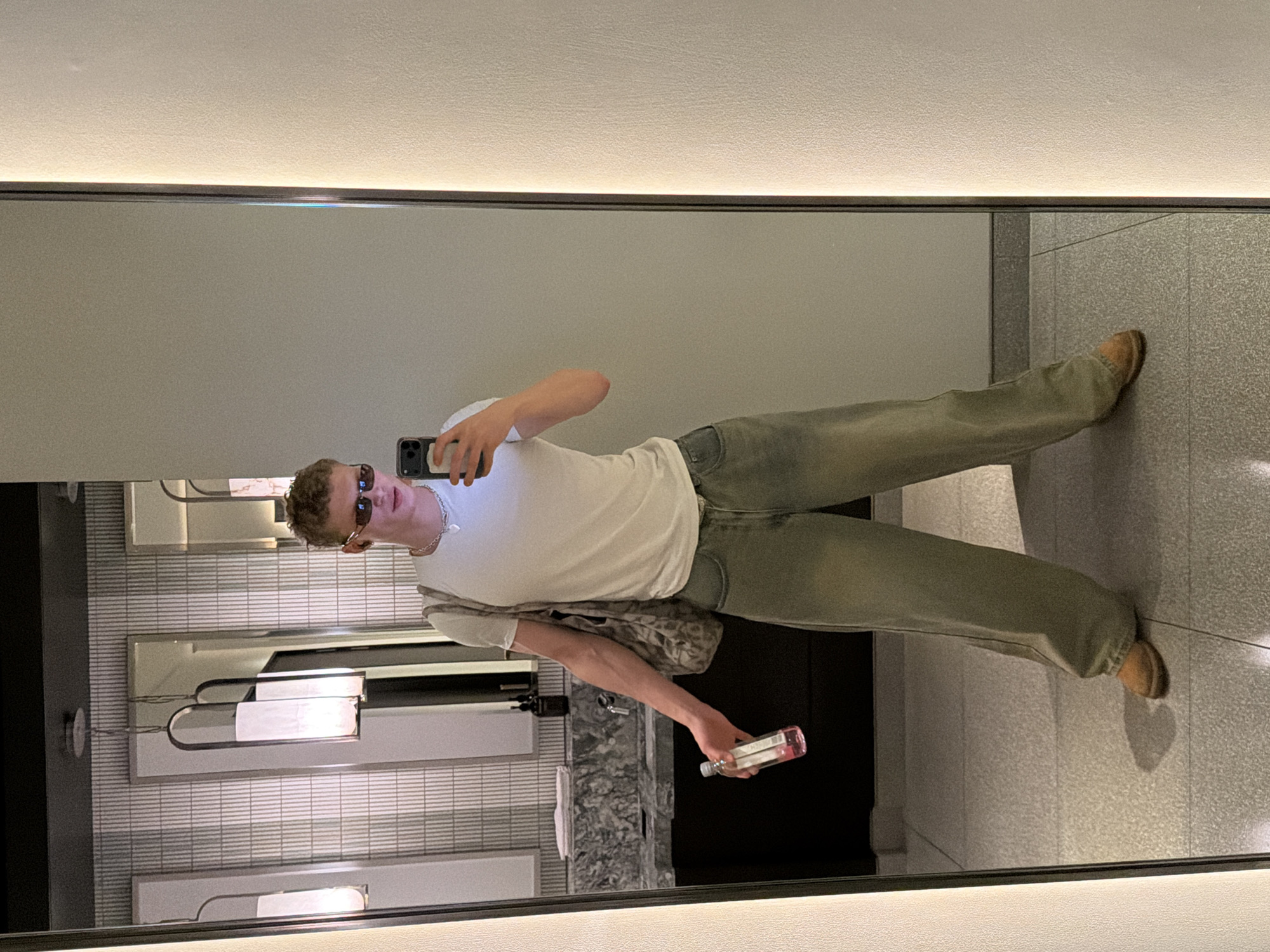A person takes a mirror selfie in a restroom. The subject is a young male with curly hair and sunglasses. He wears a white t-shirt, loose-fitting green jeans, and brown shoes. He holds a smartphone in one hand and a small bottle in the other. Behind him, the restroom features white and gray tiled walls, a marble countertop, and modern lighting fixtures. The restroom appears to be located in a modern, upscale setting. There is no specific information in the image to indicate that the location is in London, United Kingdom.