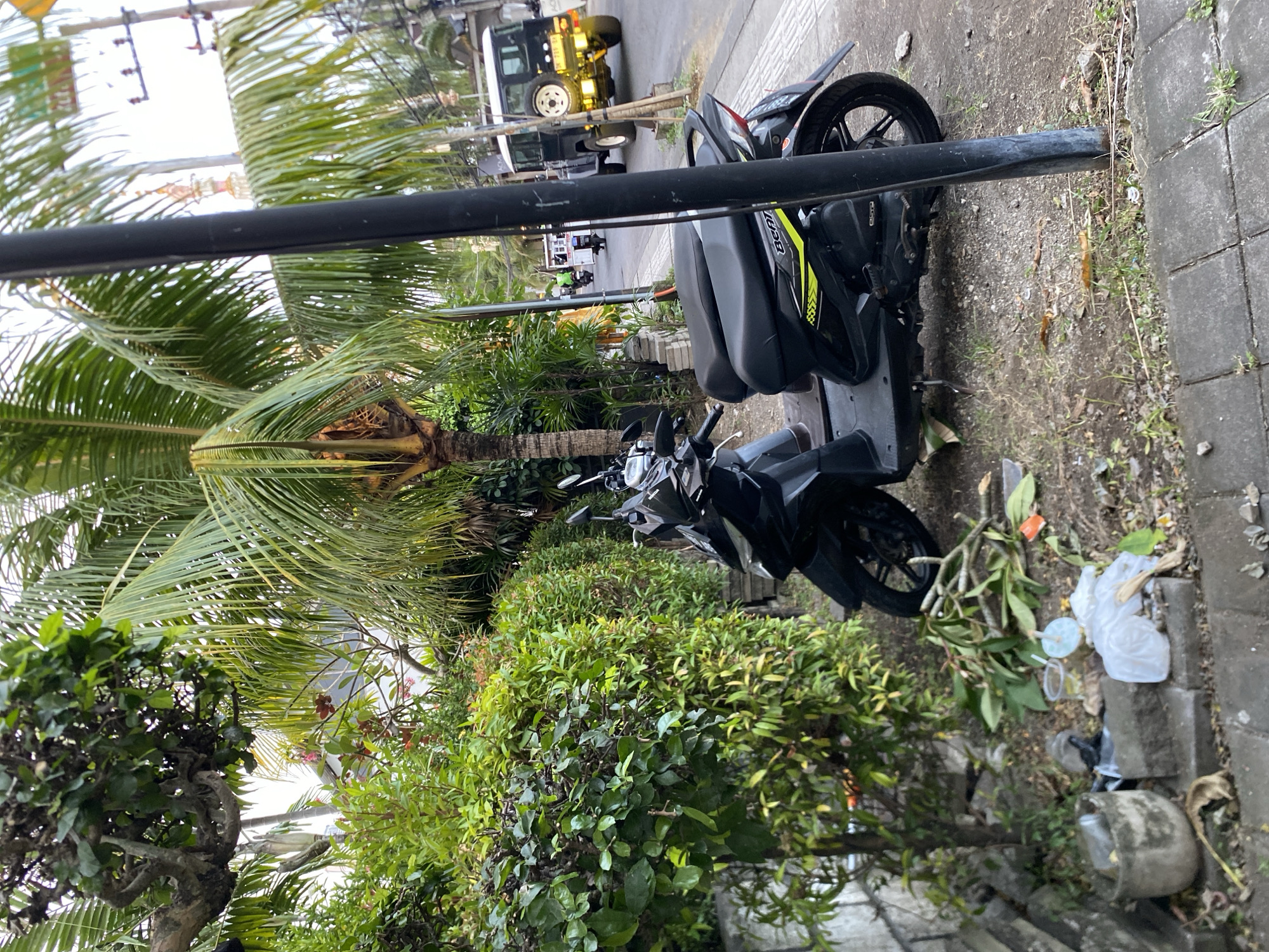 The image depicts a street scene in Mengwi, Indonesia, dominated by lush tropical vegetation and parked vehicles. In the foreground, a black and yellow scooter is parked on the side of a gray paved road, partially obscured by a dark pole. To the left of the scooter, a wall made of rough gray stones is visible, with a concrete planter containing dry leaves. Next to the planter, there is a pile of debris including plastic bags, cut branches, and a discarded cup.
The background reveals a street with traffic. A classic dark SUV with a spare tire mounted on the back is driving away from the camera. Further down the road, several other motorcycles are visible. Tall palm trees with abundant fronds line the street, interspersed with other green foliage. Electrical poles and wires are also present, indicating an urban setting. The overall impression is that of a tropical, possibly slightly unkempt, street environment.