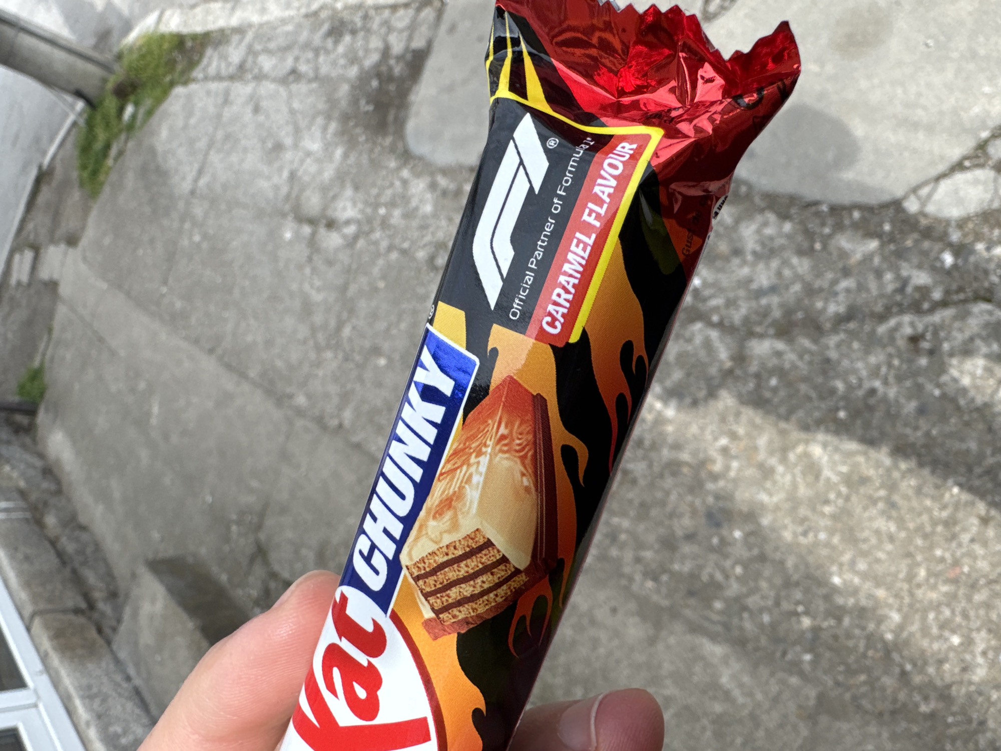 A hand holds a KitKat Chunky Caramel Flavour chocolate bar outdoors. The bar's packaging features the Formula 1 logo and a fiery design. The background consists of a concrete surface, possibly a sidewalk or curb, with some vegetation visible on the left. There is no indication this image was taken in the city of Kyustendil, Bulgaria.