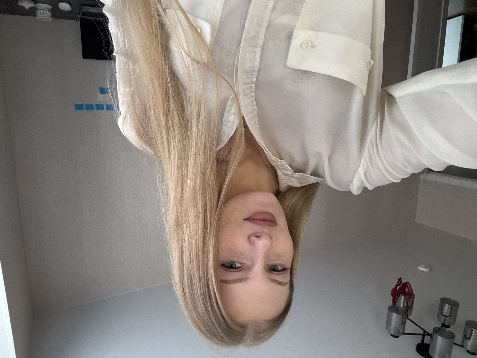 A young woman with blonde hair is taking a selfie in an indoor setting. She is wearing a white button-up shirt. Her makeup includes foundation, eyeliner, and lipstick. The background shows a ceiling with a light fixture, a wall with some blue sticky notes and a laptop on a desk. The setting does not provide any specific clues to indicate it is located in the city of Perm, Russia.