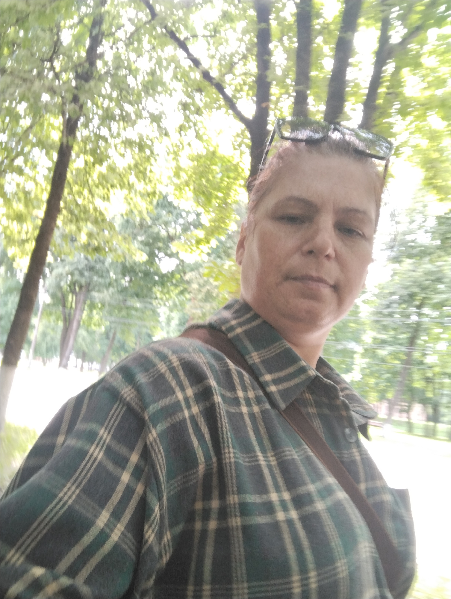 The image shows a selfie of a woman with reddish hair, wearing a green and beige plaid shirt and sunglasses perched on her head. She is outdoors, with a background of trees and foliage that appears to be in a park-like setting. The lighting suggests it is daytime, possibly overcast or with dappled sunlight. The trees are tall and leafy, and the overall atmosphere is natural and green. There is no visible indication of the city of Kingisepp, Russia, within the image itself.