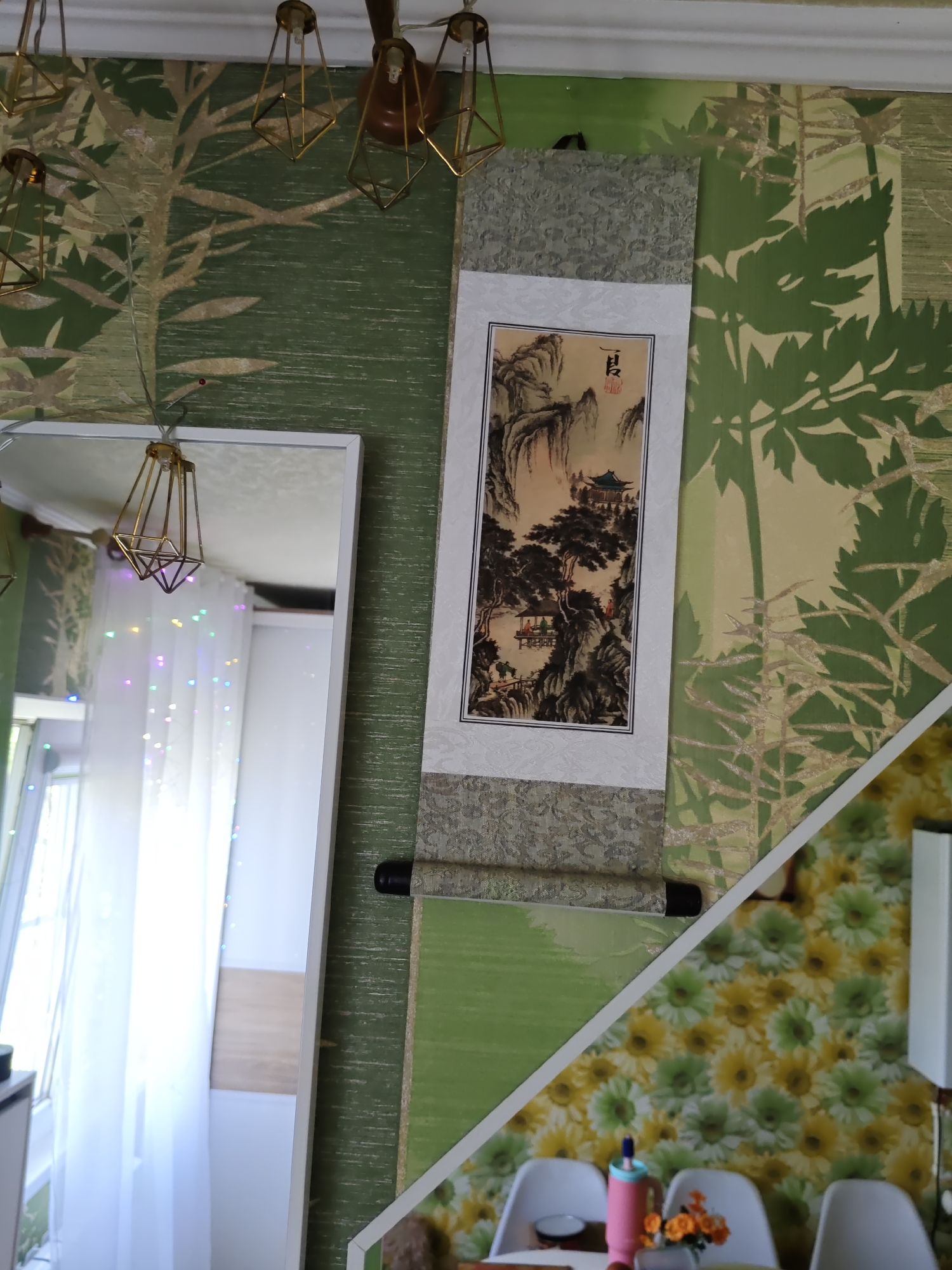 The image displays an indoor scene, likely a living space, featuring a wall decorated with a patterned wallpaper. The wallpaper on the left side exhibits a green tree motif, while the lower right section features a vibrant yellow and green floral pattern. Suspended on the wall is a scroll painting with an Asian landscape depicted. Several modern pendant lights with a geometric, gold-colored frame hang from the ceiling. A large white-framed mirror is positioned on the left side of the image, reflecting a window with sheer white curtains and colorful string lights. In the foreground, part of a dining table is visible with several white chairs, a pink water bottle, and a small vase of orange flowers. The overall impression is of a decorated interior with artistic and decorative elements. There is no indication of the city of Rostov-on-Don, Russia, or any specific geographical location.