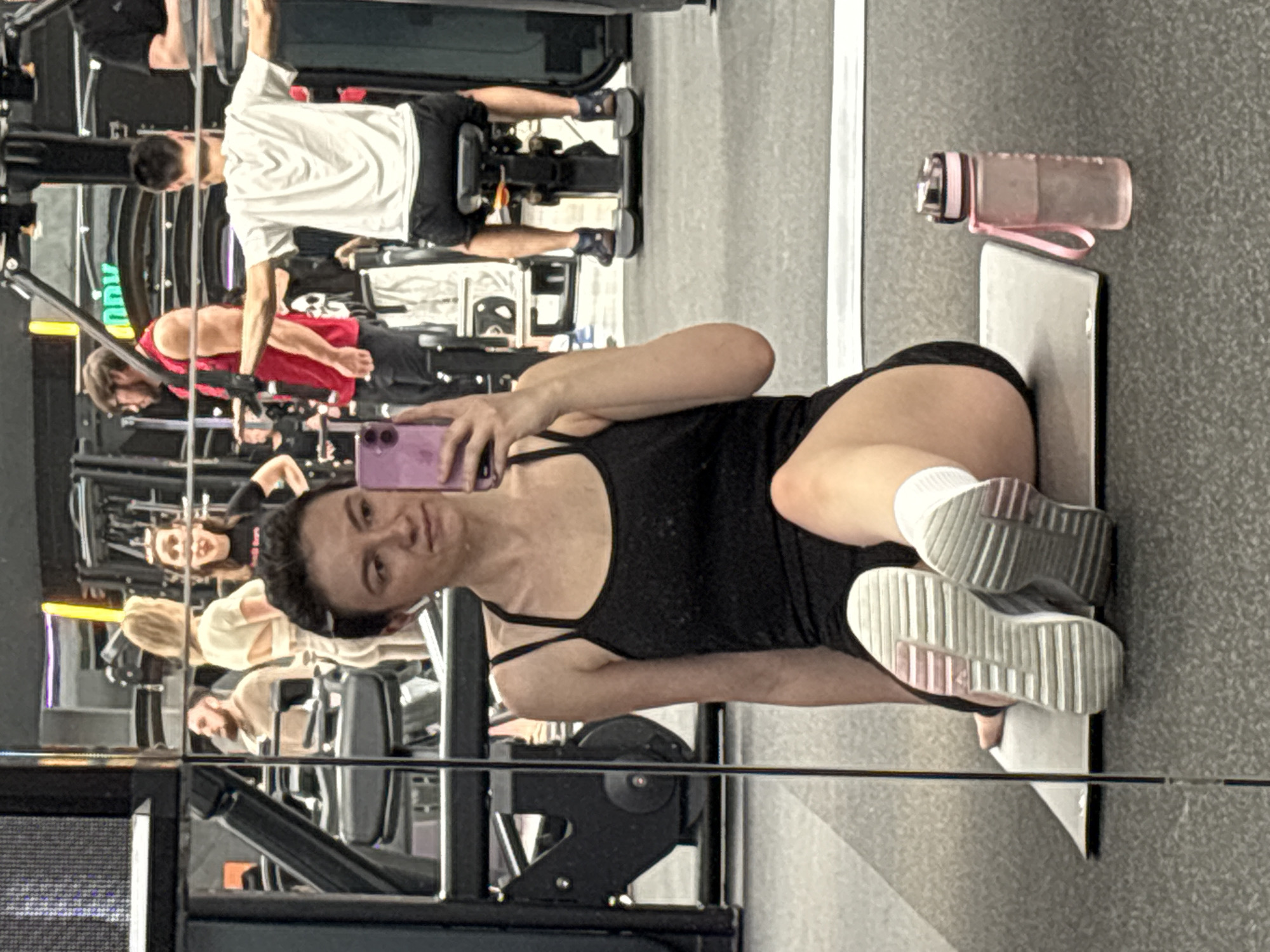 A woman in a black exercise suit lies on a yoga mat in what appears to be a gym. She is holding a smartphone, seemingly taking a selfie. She is wearing white socks and grey sneakers. A water bottle sits next to the mat. The gym setting includes workout equipment and other individuals are visible in the background, reflected in the gym's mirrors. The exact location within Kazan, Russia, cannot be determined from the image.