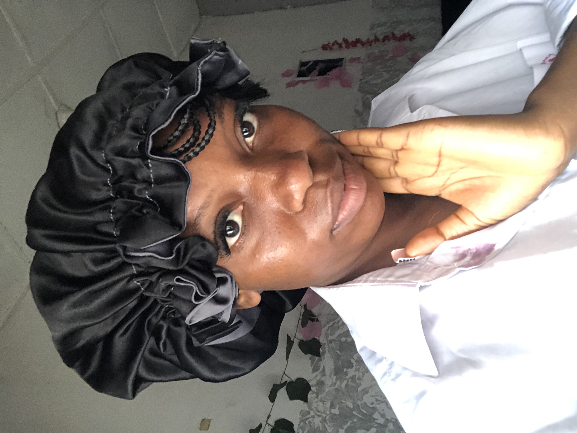 The image shows a close-up of a Black woman's face and upper body, taken from a slightly upward angle. She is wearing a black satin bonnet that covers her hair, with some braided hair peeking out. Her skin tone is medium-dark, and she has dark brown eyes and lips. She is looking directly at the camera with a neutral expression. Her right hand is resting on her face, near her cheek and eye. She is wearing a white collared shirt. There are some faint pink stains on the collar of her shirt, possibly from makeup.
The background is out of focus and appears to be an indoor setting. The wall behind her is textured and appears to be painted a light color. There is some patterned wallpaper with floral motifs visible in the background. The lighting suggests it could be daytime, but it's difficult to determine the exact time. There are no specific objects or clues that pinpoint the exact location, but the context suggests it is Nigeria. The overall mood is relaxed and casual. There is no visible text in the image.