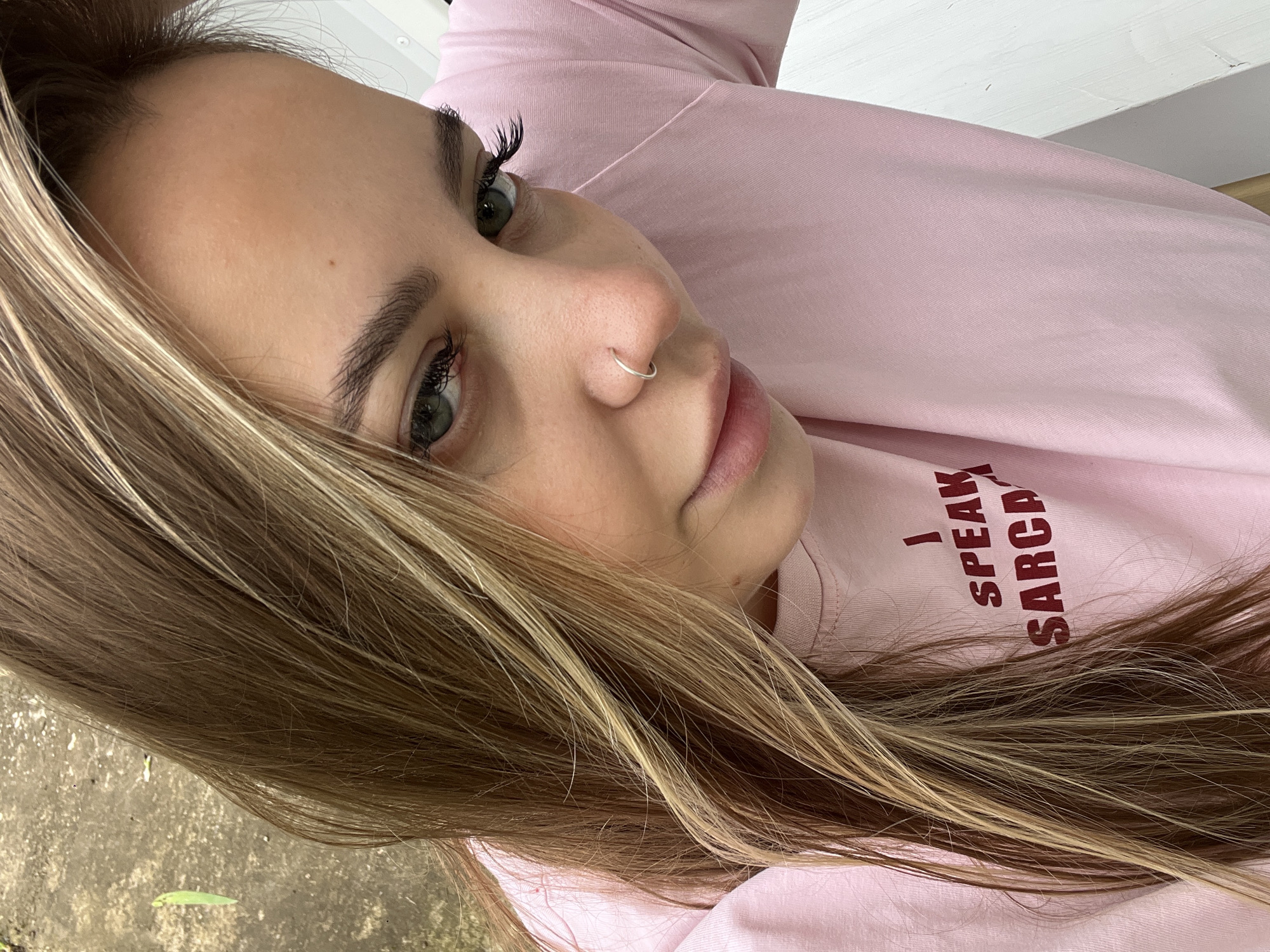 A close-up, angled portrait shot of a young woman with light brown hair and blonde highlights. She is wearing a light pink sweatshirt or t-shirt with the phrase "I SPEAK SARCASM" printed in dark red lettering. She has green eyes, defined eyebrows, long eyelashes, a silver nose ring, and fair skin. The background is blurry, with hints of greenery at the bottom left corner, possibly indicating an outdoor location. The image does not provide enough detail to place the location within Novopokrovskaya, Russia, or even confirm if it was taken there.