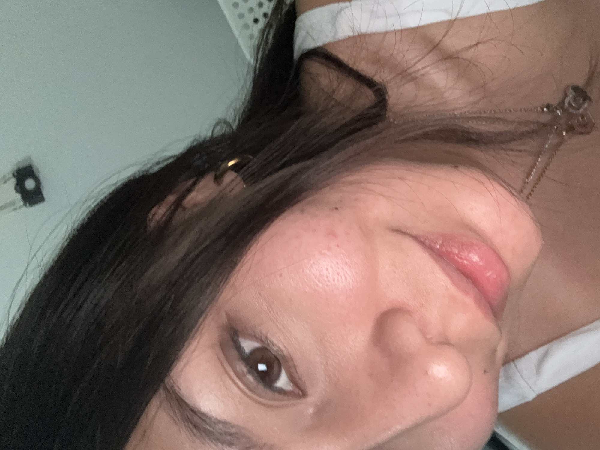 A close-up, slightly angled selfie of a woman with dark brown hair, brown eyes, and light skin. She is wearing a white top, and a delicate silver necklace. An earring is visible in her ear. The background is blurry and light-colored. No information suggests the location.