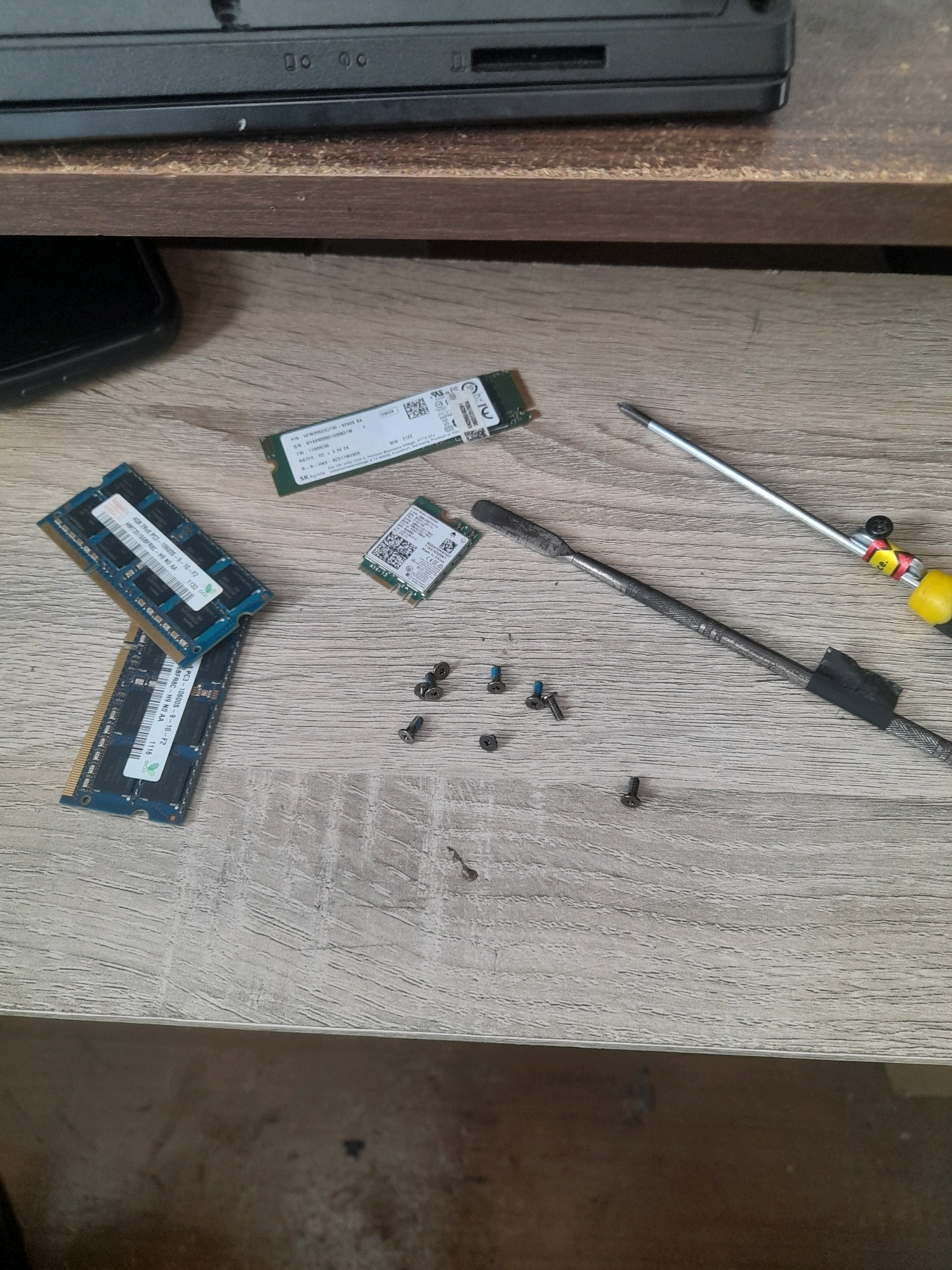 The image shows a close-up, overhead view of a wooden desk surface, likely in a workshop or personal workspace. Scattered on the desk are computer components and tools. Two RAM modules, a small M.2 solid-state drive (SSD), and a wireless card are visible. Additionally, several small screws are present, along with a screwdriver with a yellow and red handle and another tool that appears to be a pry bar or spudger, taped at one end. The background shows the edge of a black electronic device, possibly a laptop, placed on a wooden shelf. The lighting suggests an indoor environment. The image does not contain any elements that indicate a specific location in Surabaya, Indonesia, or any individuals.