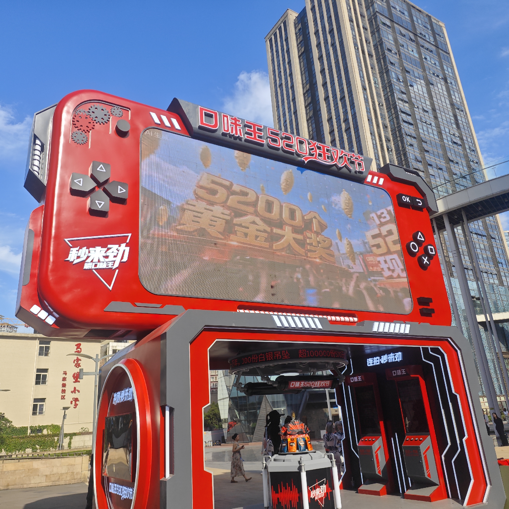 A low-angle, wide shot shows a video game-themed advertisement display, with a modern skyscraper in the background, in Yuzhong District, China.
The advertisement display is predominantly red and gray, designed to resemble an oversized handheld gaming device. The large screen displays graphics with Chinese characters and the numbers "52001." The controls on the device, including a directional pad and buttons labeled "OK," "Back," "Triangle," "Circle," "Square," and "X," are prominent. "秒来劲" is a recurring phrase visible on the display.
The advertisement display features a booth area with screens and buttons. Several people are near this booth. A woman wearing a dress is walking in the foreground, and other individuals appear to be interacting with the display.
In the background, the modern skyscraper is tall, constructed with dark-tinted windows, and surrounded by blue sky with some clouds. A skybridge connects two buildings.