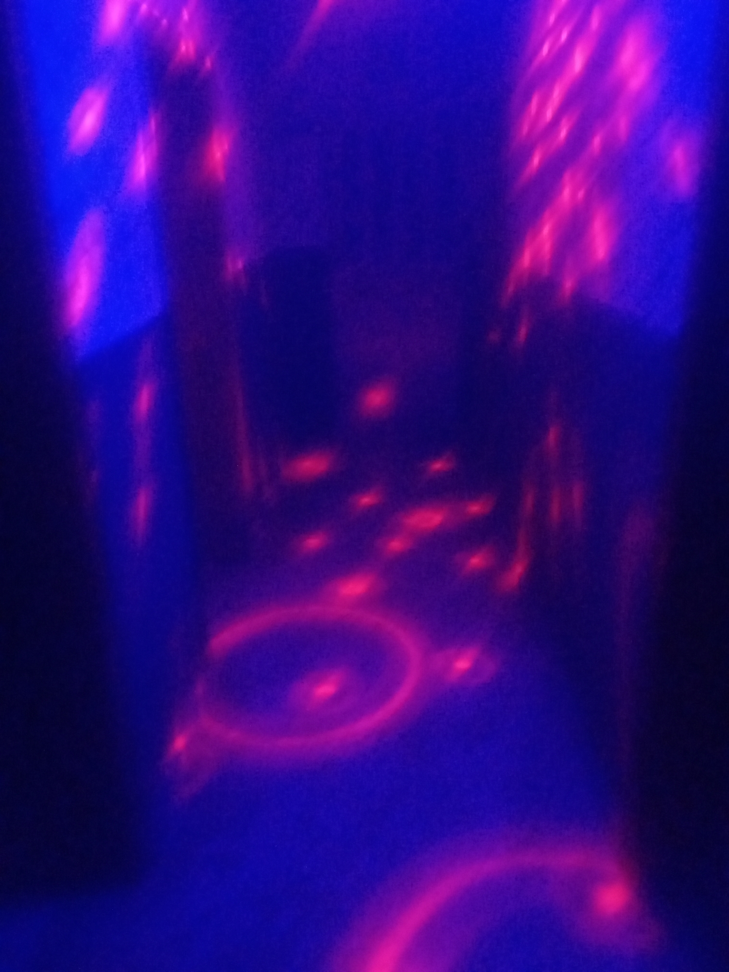 This media file depicts a dimly lit, abstract scene with dominant hues of deep blue and vibrant pink or magenta. The lighting appears to be from a source that creates blurred, luminous shapes and streaks of light against a dark background. In the foreground, there are circular patterns of pink light, one more defined than the others, suggesting a reflective surface or a projected design. Scattered throughout the image are smaller, bright pink dots or smudges of light. The overall impression is one of an indoor, possibly entertainment-focused environment, perhaps a club or a themed venue, where colored lighting is a key feature. The abstract nature of the image, with its blurred forms and intense colors, creates a somewhat disorienting yet visually striking effect. There is no discernible text, people, or specific objects clearly identifiable, making it difficult to determine the exact activity or event beyond a general sense of an atmospheric setting. The time of day or weather are not inferable. Given the provided location context of Sabon Gari Nassarawa, Nigeria, this could be a view within a local establishment utilizing this type of lighting.