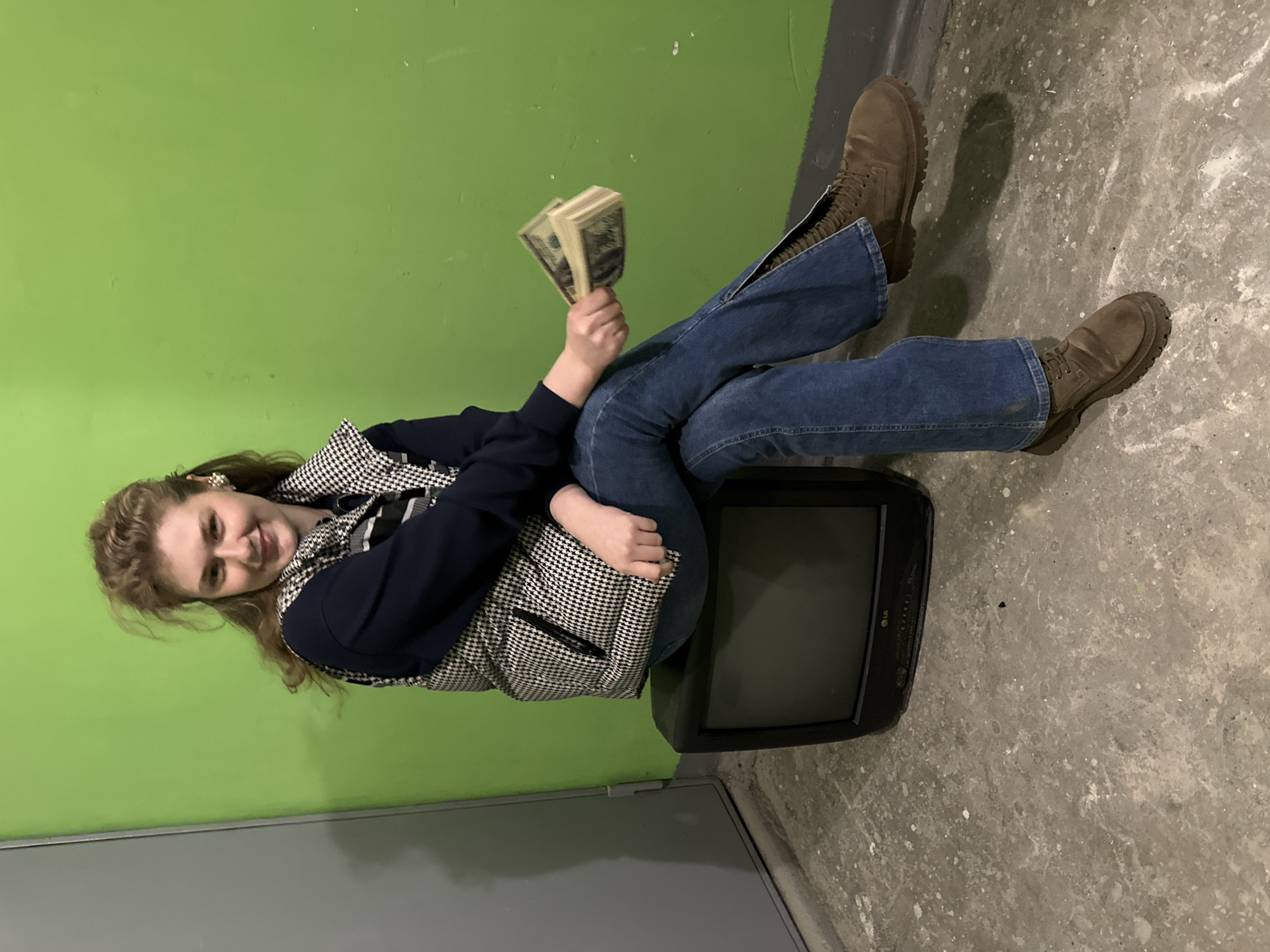 A young woman is seated on a black LG television set in a room with a green wall and a grey concrete floor. She is holding a wad of US dollar bills in her left hand. She wears blue jeans with zippers on the side, brown boots, and a dark blue sweater under a houndstooth puffer vest. The scene is indoors, possibly a storage area or basement with minimal decoration, typical for a generic, nondescript interior space. Location within the city of Kazan, Russia, cannot be determined from visual data.
