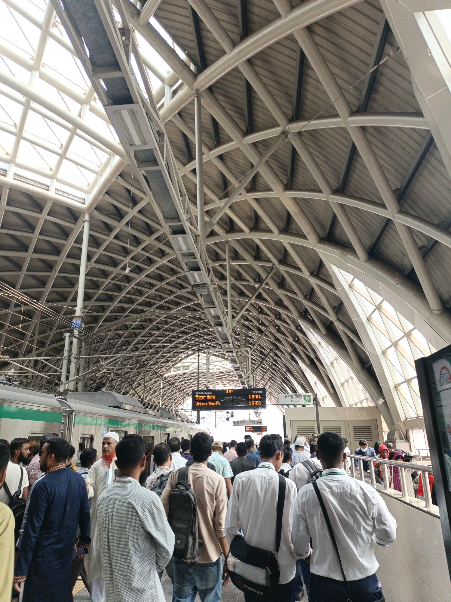 The image depicts a busy train station in Dhaka, Bangladesh. A crowd of people is gathered on the platform near a train with green and white cars. A digital display reads "Uttara North" in Bengali and English, indicating the destination. The station has a distinctive arched roof structure made of steel, allowing natural light to filter in. Some individuals wear masks, indicating a potential health concern.