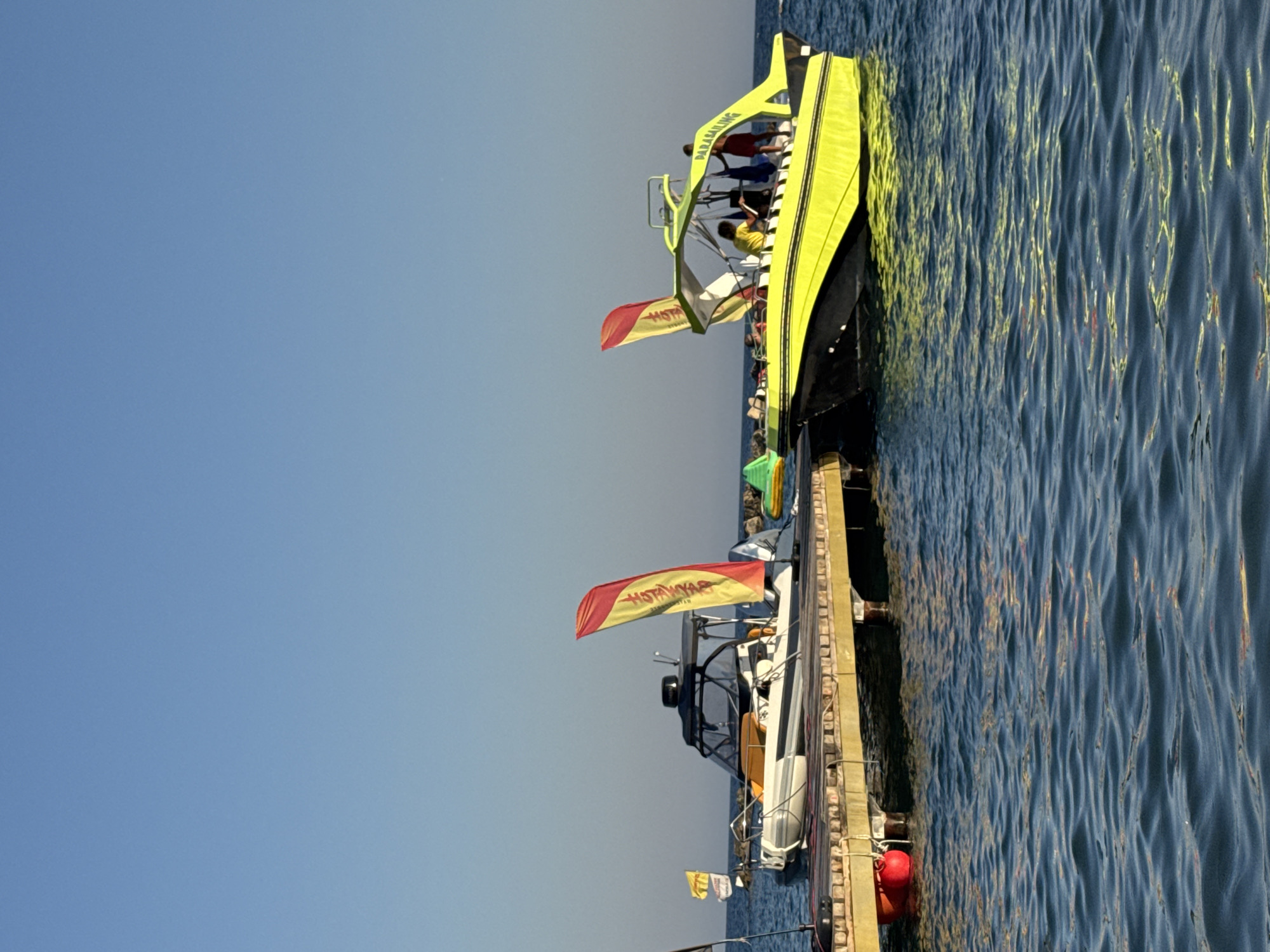 Two boats are docked side-by-side at a wooden pier on a clear day in Agios Tychona Community, Cyprus. The boat on the left is white with a dark canopy and a yellow and red flag. It is moored to the pier, with a red buoy visible beneath. The boat on the right is bright yellow with a black hull and a green canopy. Several people are on board this boat, and the word "PARASAILING" is visible on its side. The water is calm and blue, reflecting the sunlight. In the background, the sky is a clear, bright blue, with a few dark rocks visible along the shoreline.