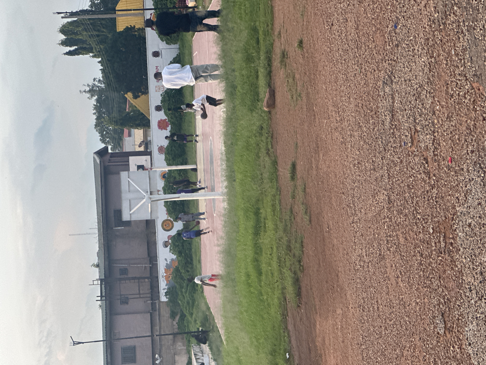 This image captures an outdoor basketball court scene in an urban or semi-urban environment in Unknown, Unknown, under an overcast sky. In the foreground, a reddish-brown dirt and gravel path occupies the lower third of the frame, transitioning to a strip of green grass that separates it from the basketball court. A small brown rock is visible on the grass strip towards the center-right.
On the basketball court, which features a faded reddish-orange surface with white court markings (including the key and center circle), multiple individuals are present. Approximately ten to twelve males, primarily appearing to be young adults or teenagers, are engaged in or observing a game of basketball. One young male in a white t-shirt and dark shorts is actively dribbling a basketball near the center of the court. Another male in a blue t-shirt and dark shorts is running on the court. Two basketball hoops with white backboards and poles are visible, one partially obscured on the left and another more central.
On the right side of the court, two adult males are standing and observing. One wears a white t-shirt and light-colored pants, while the other wears a black t-shirt and dark pants, with his back partially to the camera. On the far left of the court, a person is seated on the ground in the shade of a concrete structure.
Behind the court, a white wall runs horizontally, adorned with various murals or graffiti, including abstract orange and black shapes, circular emblems, and logos. Green bushes and foliage are growing in front of the lower section of this wall.
In the background, a large, two-story building with a reddish-brown facade and a dark green roof dominates the left-center. This building features multiple windows, some with security bars, and an external ladder-like structure on its side. A black utility pole stands in front of it to the left. On the roof of this building, a single chair is visible, along with some sparse vegetation. To the right of this building, more distant structures are visible, including one with a bright yellow roof. Numerous tall, slender trees and other foliage are scattered throughout the background. Utility lines are visible against the light grey, cloudy sky.