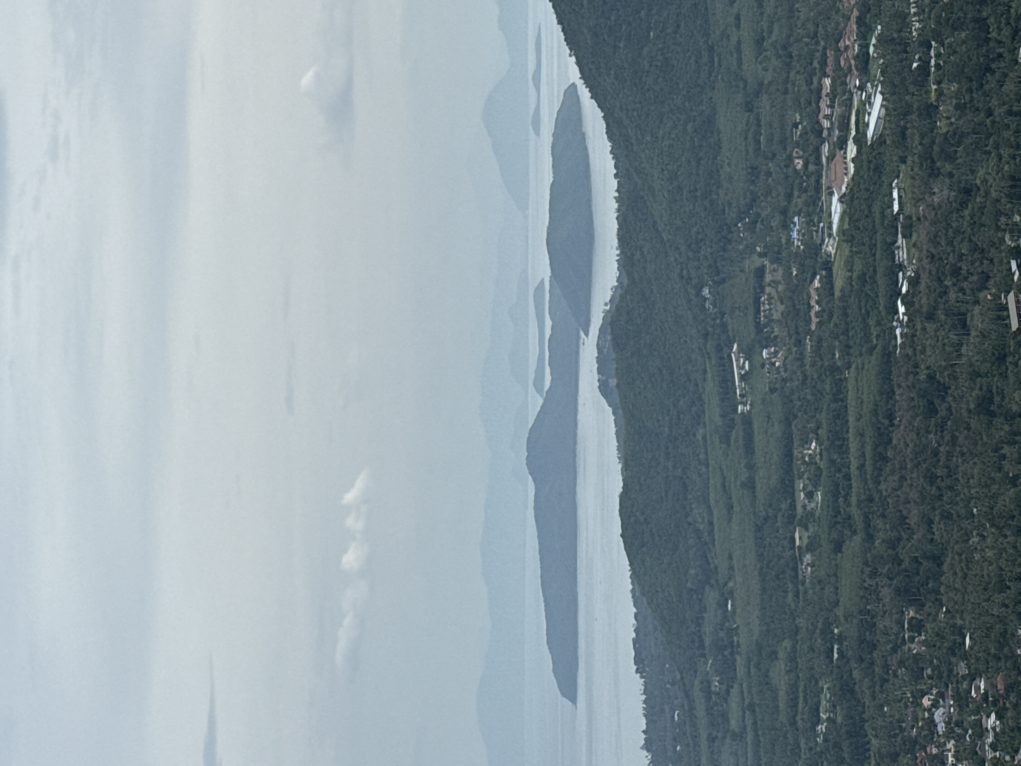 This is an aerial, high-angle view of a lush, mountainous landscape on a hazy, overcast day. In the foreground, dense green trees cover rolling hills. Scattered throughout the foliage are a few small clusters of buildings with red and white roofs, indicative of a residential or resort area.
Further into the distance, the landscape transitions to a body of water, likely the sea, dotted with several large, dark green islands of varying sizes. These islands are also covered in thick vegetation.
The horizon is blurred by haze, with layered silhouettes of more distant islands or mountain ranges visible. The sky is a pale, muted gray with diffuse white clouds. The overall impression is one of a tropical, secluded natural environment. This scene is consistent with the topography of Ko Samui District, Thailand, known for its mountainous terrain, tropical forests, and numerous offshore islands.