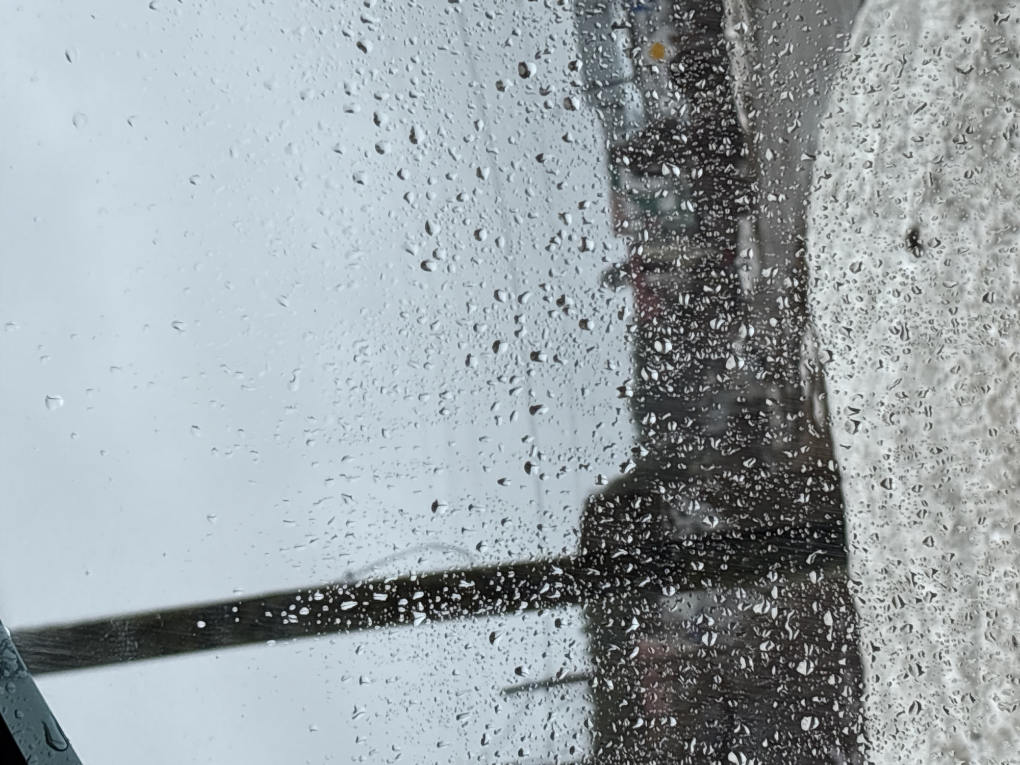 The image, taken in what appears to be rainy or snowy weather, shows a view through a wet window. Numerous water droplets or snowflakes are visible on the glass surface, partially obscuring the scene beyond. The exterior scene appears to be urban, with blurry shapes suggesting buildings or infrastructure. A dark, possibly metallic, rail or bar runs horizontally across the lower portion of the window. A large white area to the right may be snow or ice accumulation on a nearby surface.
While the image is taken in Прикамский, Russia, it is difficult to know what subjects, objects, actions, interactions, and the environment are without a clear view of the scene.