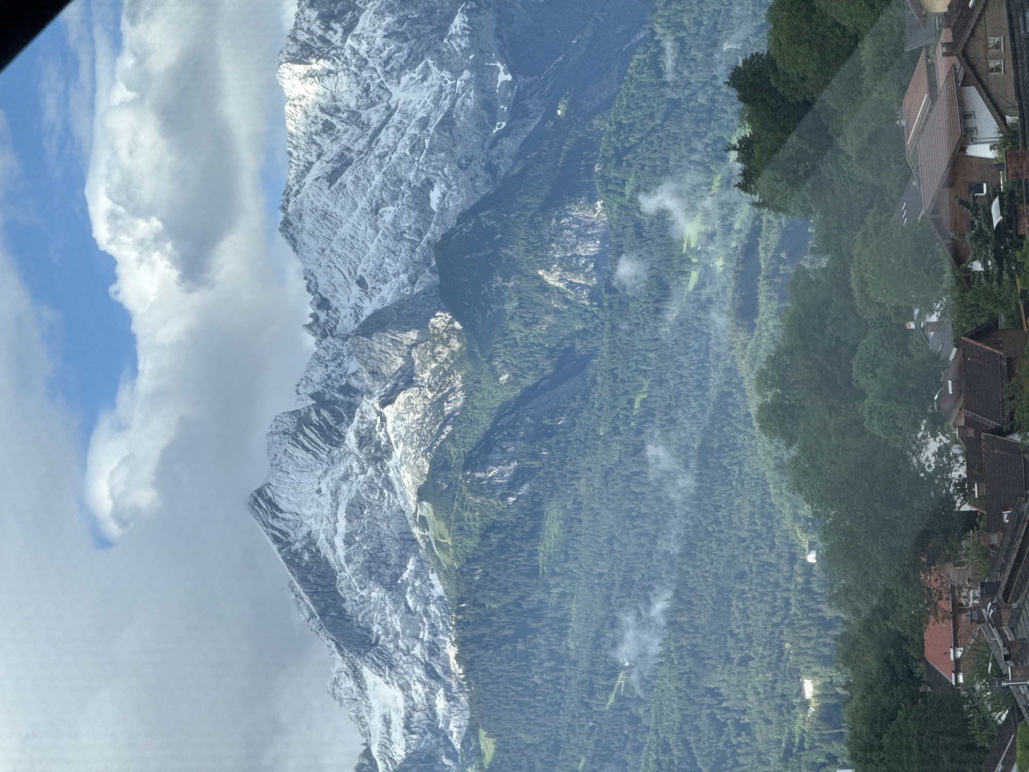 The image shows a panoramic view of the Zugspitze mountain range, with its peaks dusted with snow, overlooking the town of Garmisch-Partenkirchen, Germany. The lower slopes of the mountains are covered in lush green forests, with some patches of mist clinging to the trees. In the foreground, the rooftops of houses in Garmisch-Partenkirchen are visible, with red-tiled roofs and chimney stacks dotting the landscape. The sky above is a mix of blue and white clouds, with the sun occasionally breaking through, illuminating parts of the mountain. The overall scene is one of natural beauty and tranquility, with the majestic mountains providing a stunning backdrop to the charming town.
