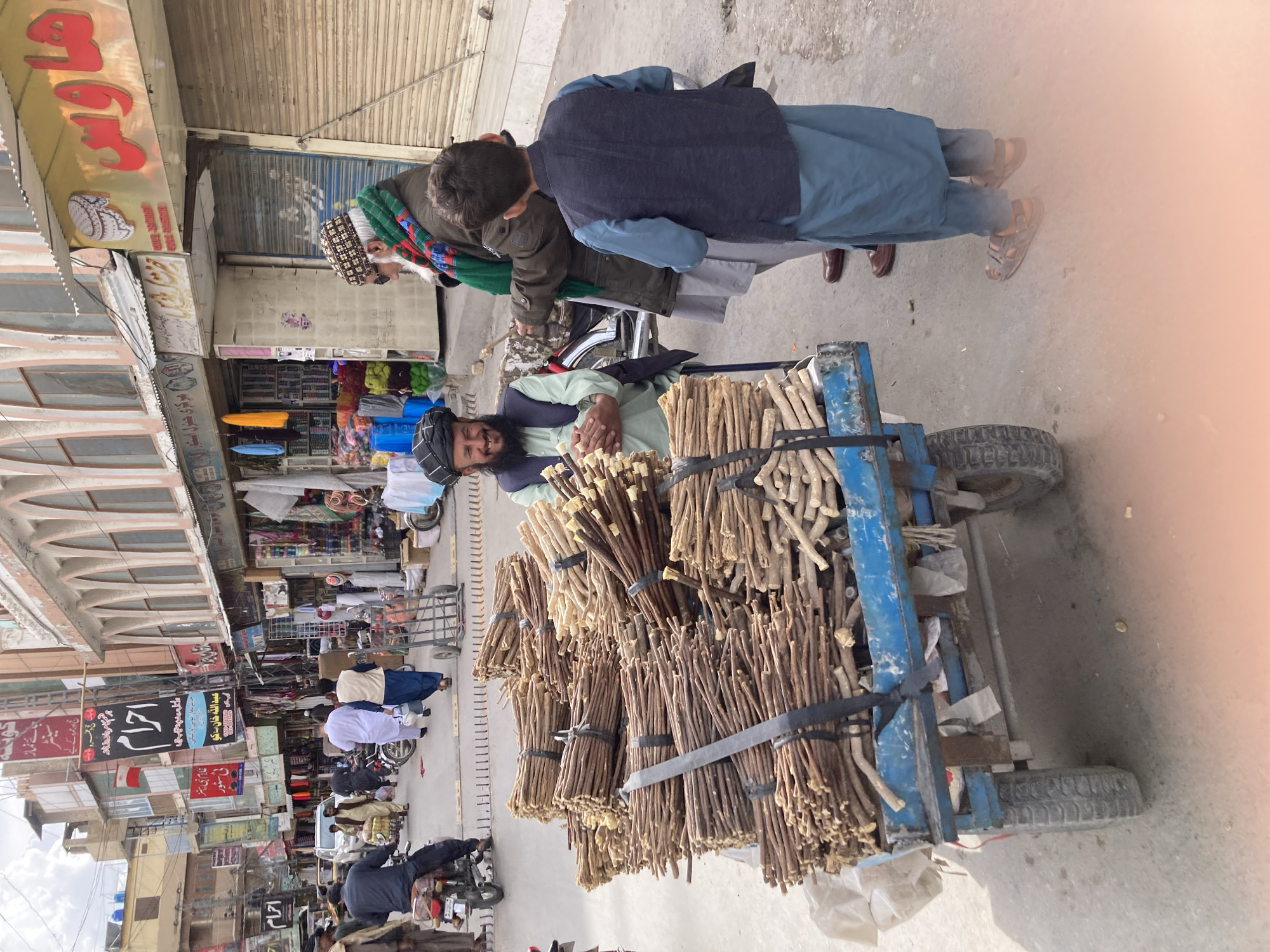 Here is a description of the image.
**Scene:** A bustling street scene in Quetta City Tehsil, Pakistan, filled with shops and people.
**Foreground:**
A man with a turban and beard sits on a blue handcart laden with bundles of what appears to be sticks, possibly "miswak" (teeth-cleaning sticks). He smiles slightly. Two people (possibly customers) are standing to the right, examining his wares.
**Background:**
The street is lined with buildings and shops. Signs with Urdu or Arabic script adorn the buildings. The shops seem to offer a variety of goods. Many other pedestrians and motorized vehicles (motorcycles) are present, suggesting a busy commercial area.
**Overall Impression:** The photo captures a typical street scene in a South Asian urban area, with a street vendor selling goods to passersby.