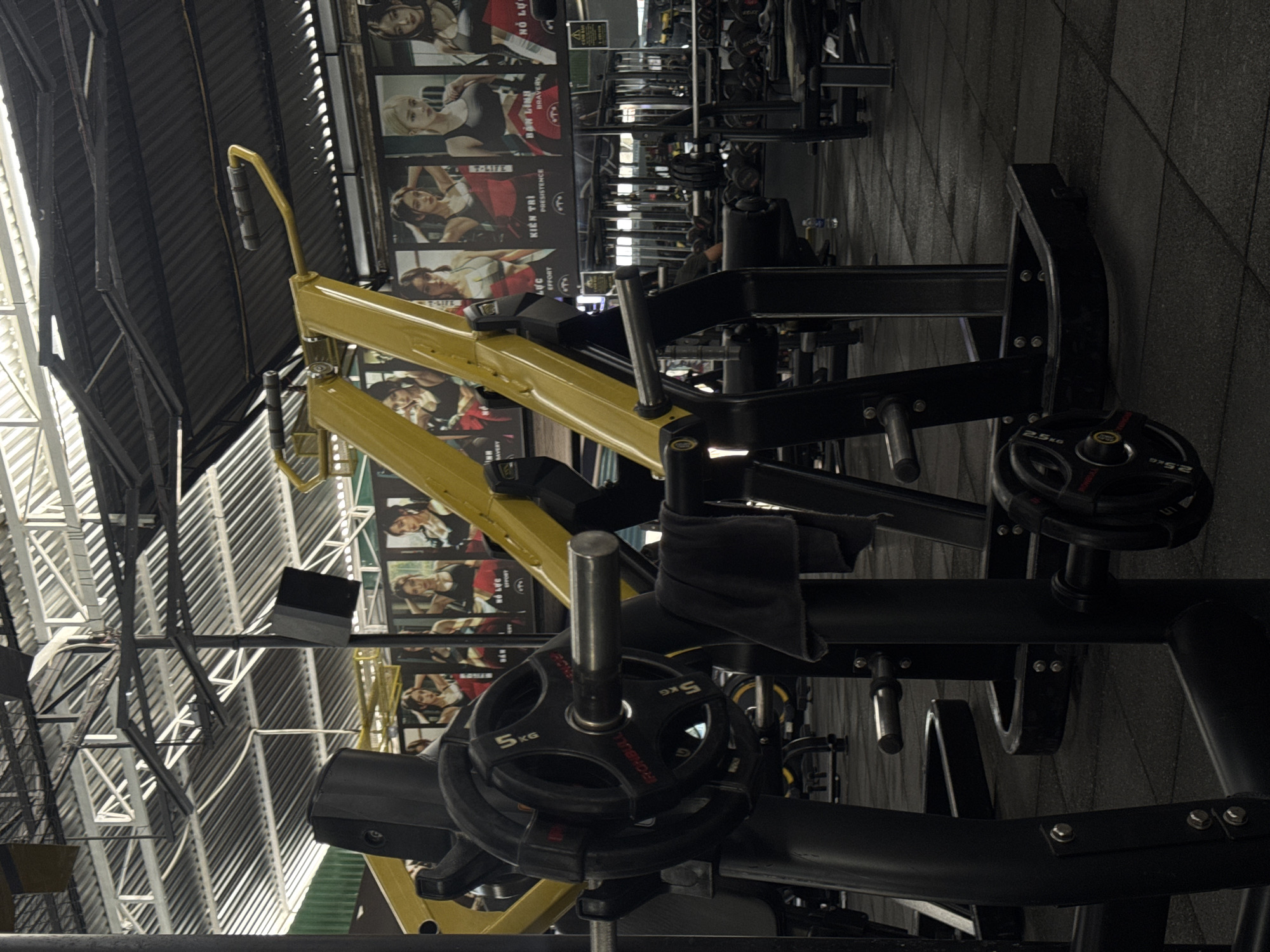 The image captures a detailed view of a gym interior, focusing on exercise equipment and decorative posters. In the foreground, a black weightlifting machine is prominently featured. A barbell with multiple 5kg weight plates is loaded onto the machine. A black towel is draped over the bar, and a portion of another weight plate, marked 2.5 KG, is visible on the right side of the frame.
Behind the weightlifting machine, several large posters are affixed to the wall, each depicting a young woman in athletic wear. The posters appear to be motivational or promotional in nature, with Vietnamese text present on some. The gym environment is characterized by a dark, textured floor, likely rubberized for shock absorption. Overhead, a metal roof structure with exposed beams and corrugated roofing is visible, along with industrial lighting fixtures. In the background, additional gym equipment, including weight racks filled with dumbbells and other machines, can be seen. A person is partially visible in the middle ground, seated at a machine. The overall atmosphere suggests an active and well-equipped fitness facility.