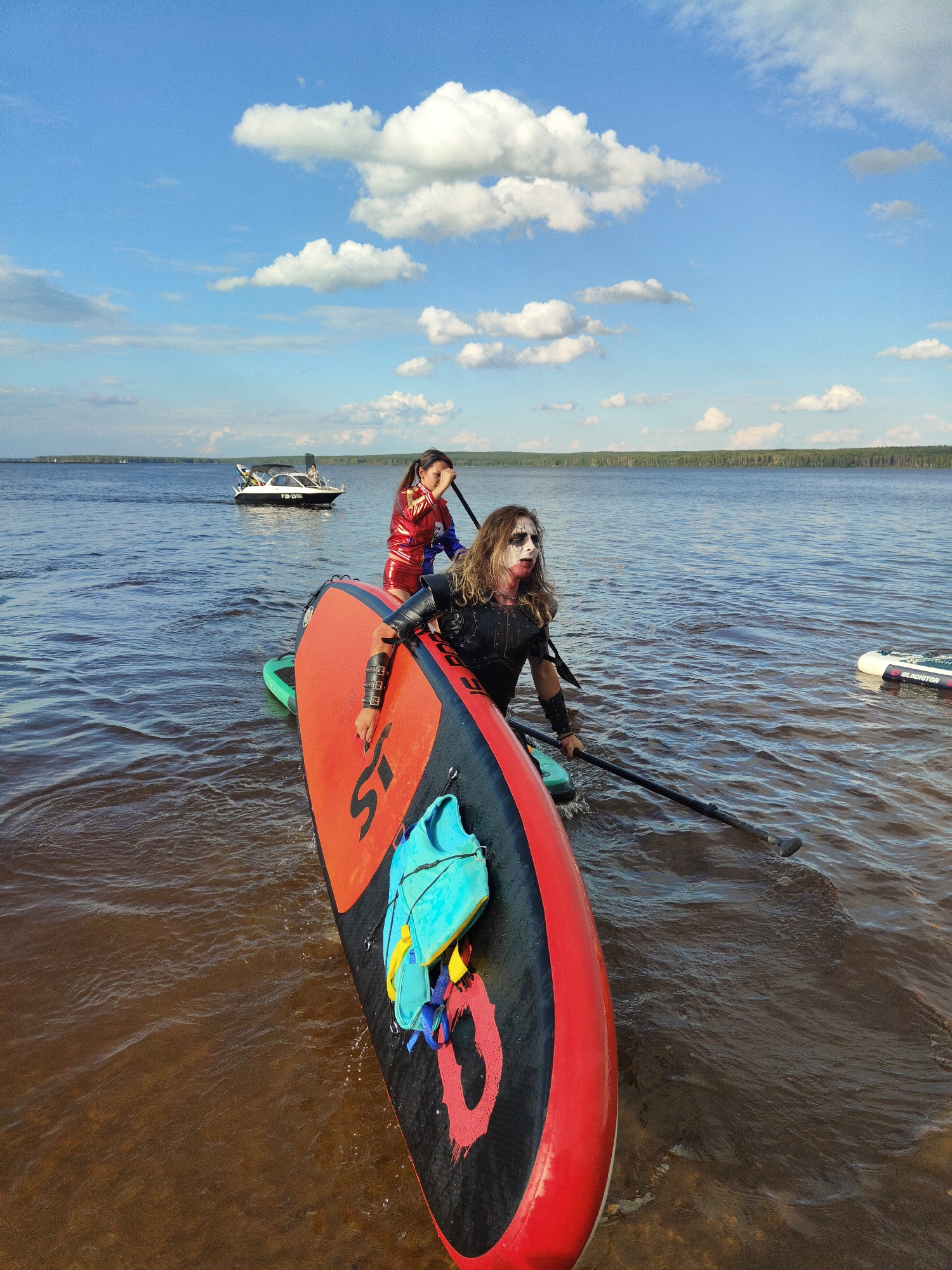 Two people are paddleboarding on a body of water. The person in the foreground is wearing dark clothing and has dramatic white and black face paint, resembling a punk or gothic style. They are holding a paddle and appear to be in motion. Behind them, another person is also paddleboarding, wearing a red and blue jacket and shorts. A white motorboat is visible in the background on the water. The scene appears to be a recreational outing on a lake or river under a partly cloudy blue sky, with a treeline visible on the distant horizon. The water is brown and somewhat choppy. A life vest is attached to the front of the paddleboard in the foreground.