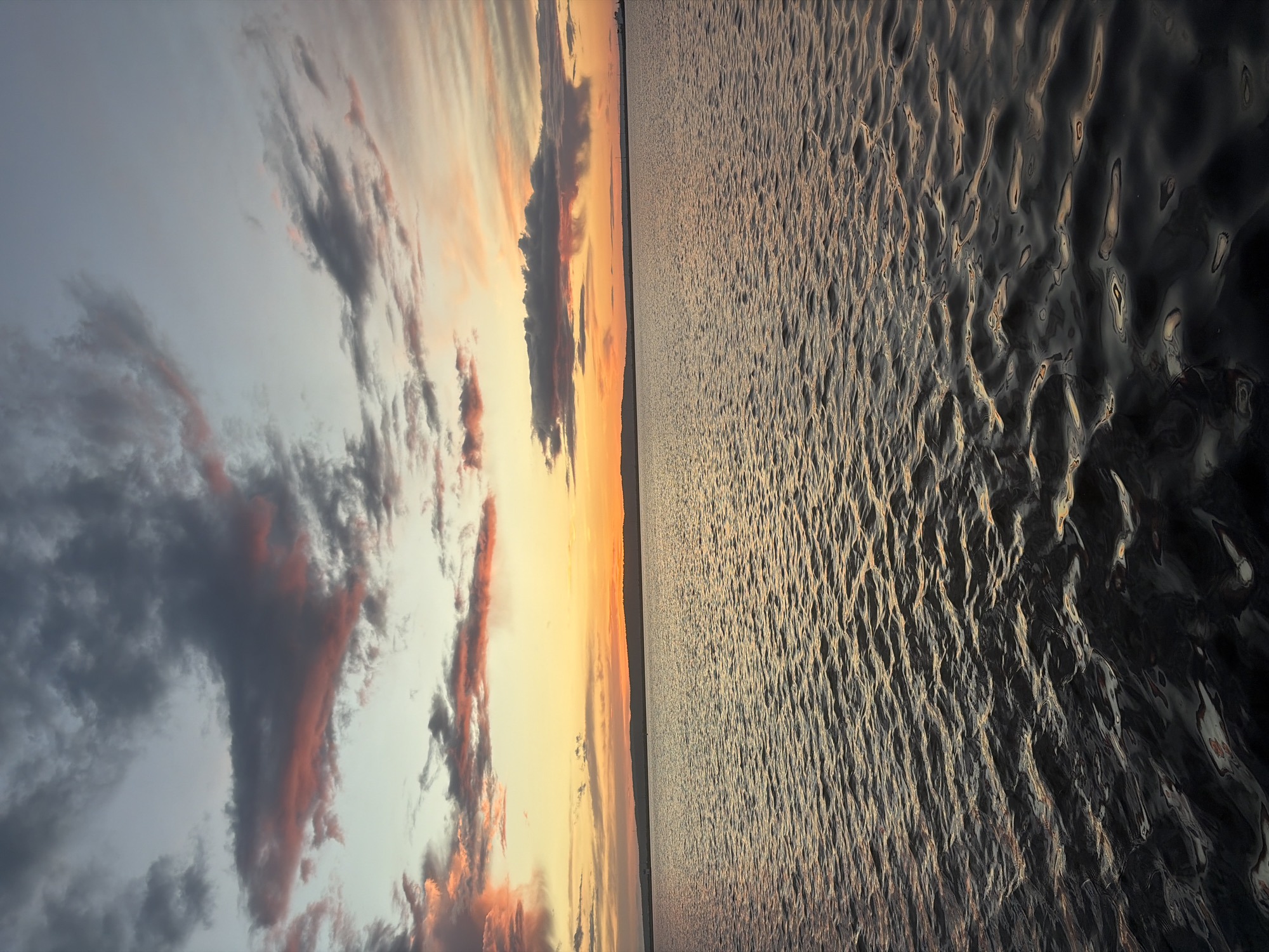 The image shows a sunset over water. The sky is filled with clouds, some tinted with orange and pink from the setting sun. The water is rippled and reflects the light from the sky. The location appears to be in the city of Unknown, Unknown.
