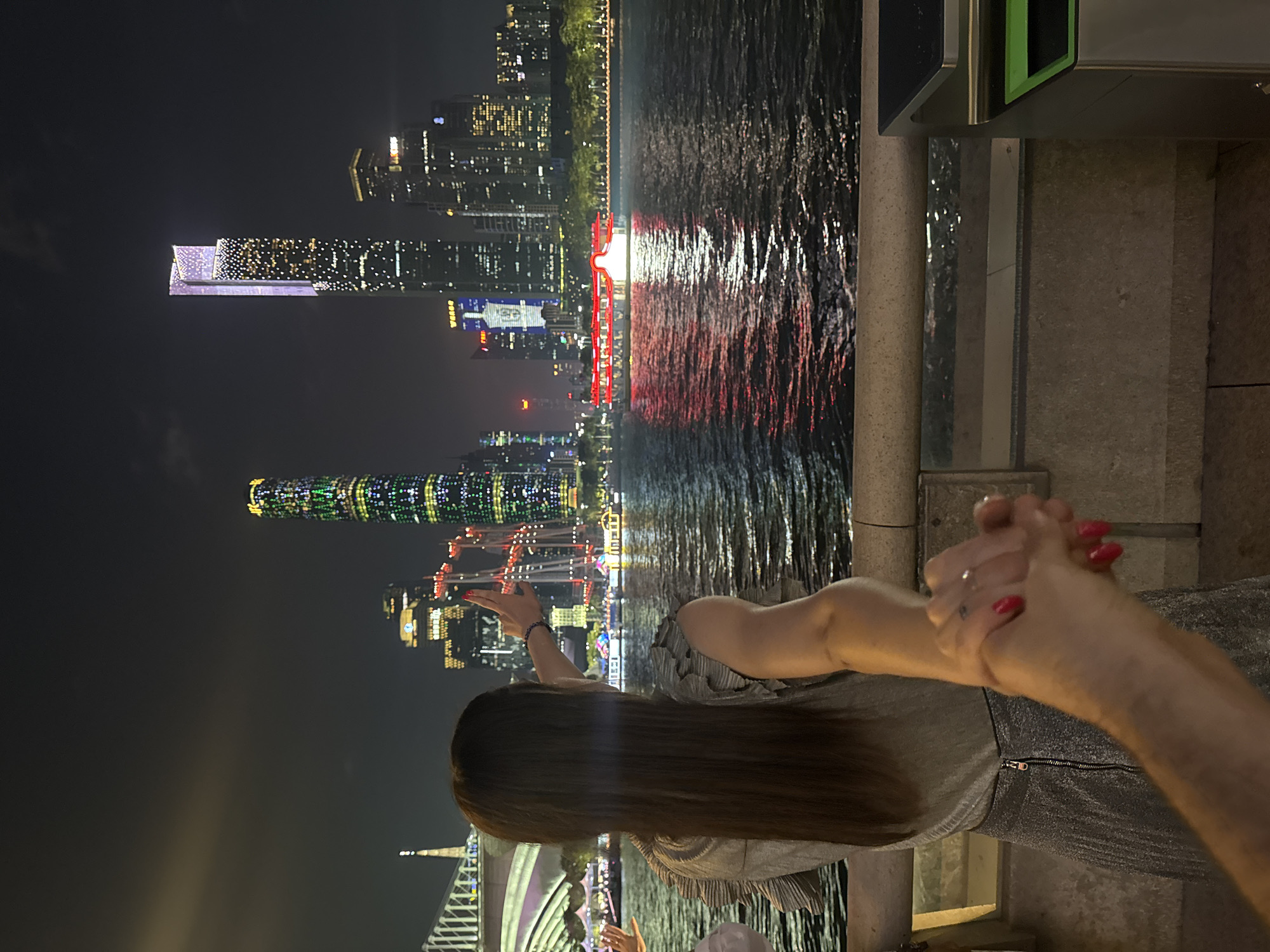 A nighttime photograph shows a woman, likely in the Tianhe District of Guangzhou, China, in front of a river with a cityscape lit up in the background. The woman is seen from the back, her long hair falling down her back. She is wearing a grey top with silver glitter. Her left arm is outstretched toward the cityscape. She is holding hands with a person to her right; only their arm and hand are visible. The fingertips are painted red.
The backdrop features a body of water reflecting lights from the buildings along the river. Buildings of varying heights are visible, including one tall skyscraper lit up with various colors and patterns, and another with white and yellow lights. In the center, there appears to be a red structure, possibly a boat or part of the cityscape decoration. The sky is dark, indicative of nighttime.