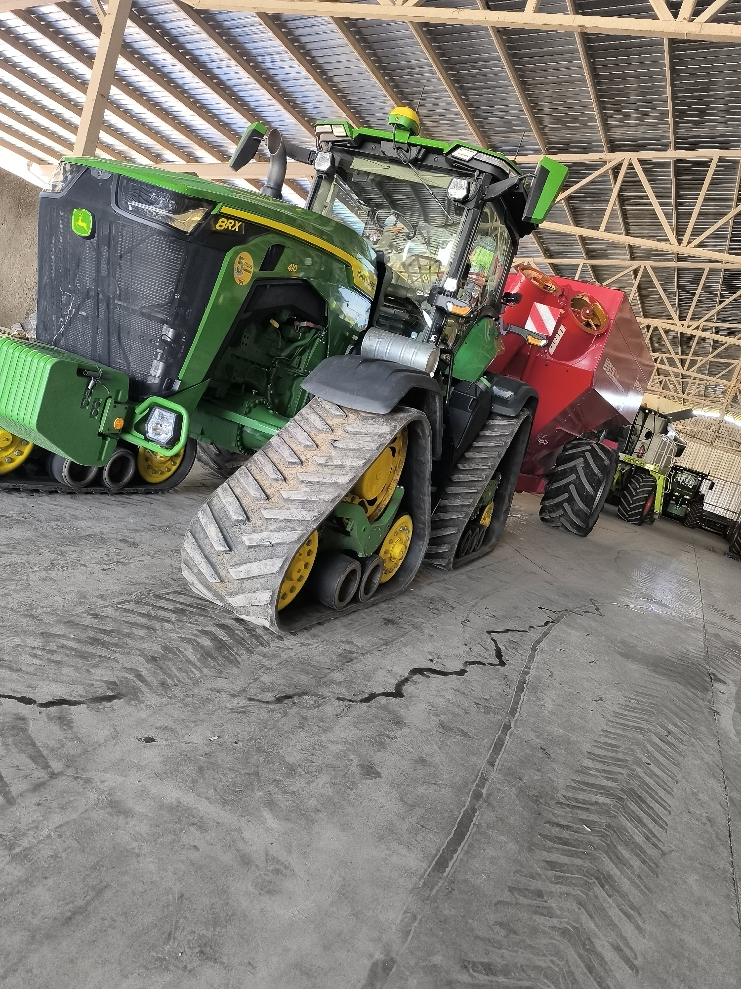 The image shows a John Deere 8RX tractor parked inside a large agricultural building. The tractor is green and yellow and features tracks instead of wheels. To the right of the tractor, a red agricultural implement is attached. Further in the background, another green tractor and a combine harvester are visible. The floor of the building is concrete, and the ceiling is made of corrugated metal panels supported by wooden beams. The overall scene suggests an agricultural setting in Maior Uzunovo, Bulgaria.