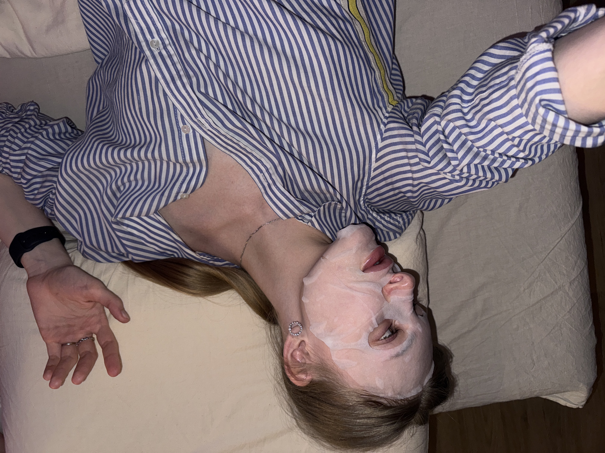 A woman lies on a beige pillow or cushion wearing a white facial mask. She is dressed in a long-sleeved blue and white striped shirt that is slightly unbuttoned at the top. A thin silver necklace is visible around her neck. Her left wrist has a black band, and a ring is on her finger. She is wearing a circular earring. The environment appears to be indoors with the pillow or cushion lying on a surface that is a neutral tone. The image does not provide information that can be used to infer the city of Perm, Russia.