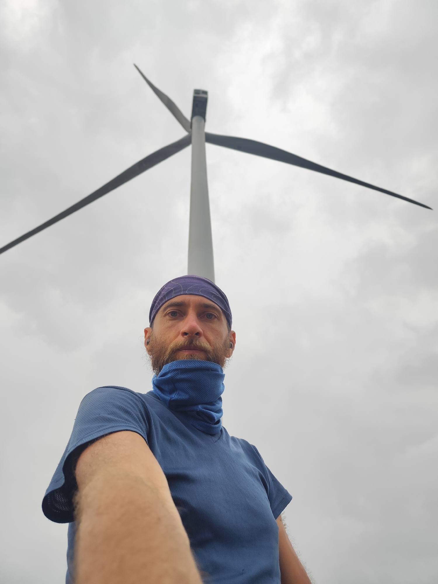 A low-angle, medium-shot photograph features a man in the foreground with a wind turbine in the background under a cloudy sky.
The man has fair skin, a red beard, and blue eyes. He wears a blue head covering and a blue mesh neck gaiter pulled up to his lower nose. He wears a short-sleeved blue shirt. The man is holding the camera, his arm extending into the frame.
Behind the man, a large, grey wind turbine stretches into the cloudy sky. The turbine has three blades.
Based on geolocation data, this photograph was taken near Trường Thọ in Vietnam.