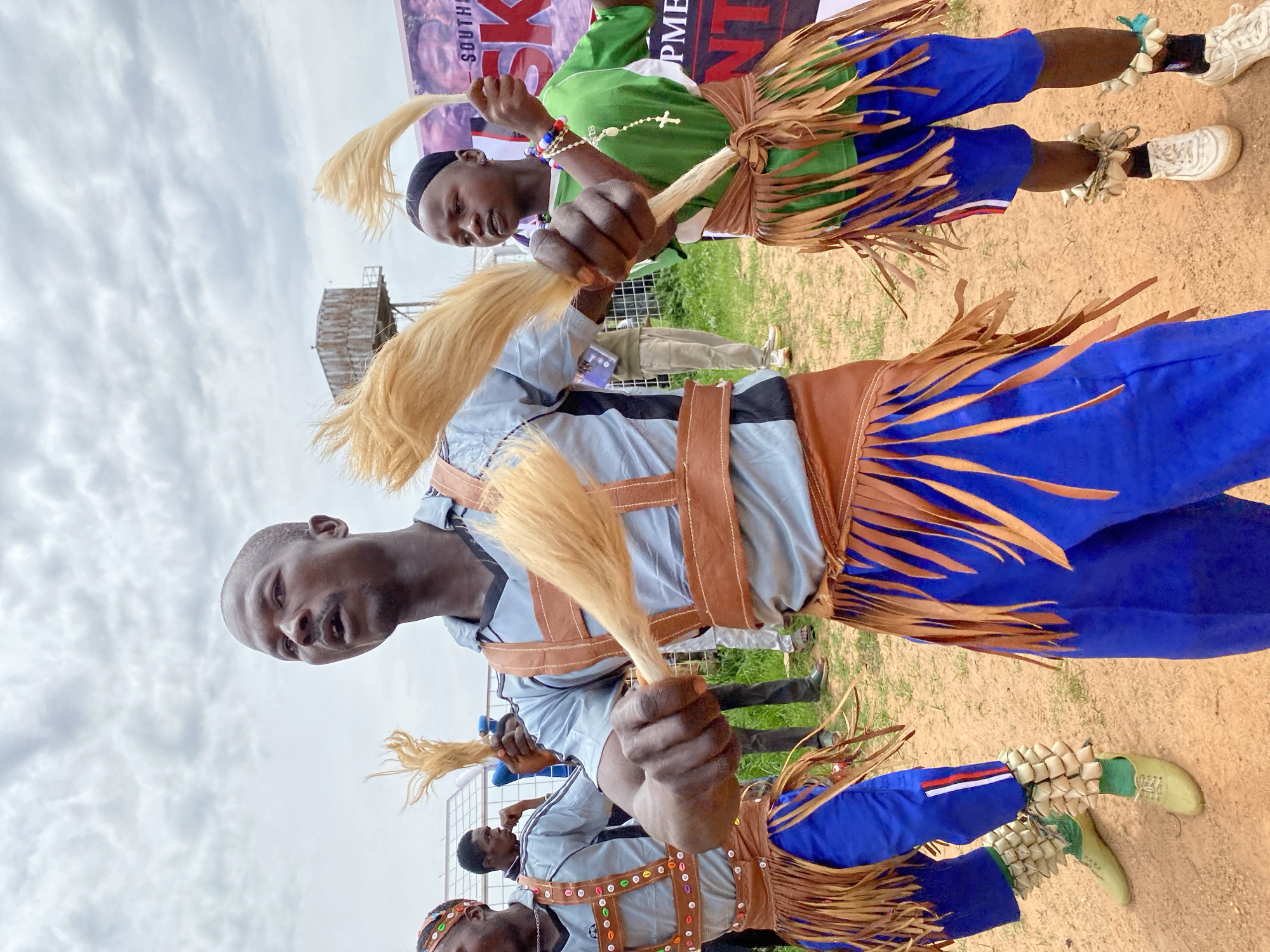 In an outdoor setting, two men dressed in ceremonial attire are positioned close to the camera. The man closest to the viewer is wearing a light blue, long-sleeved shirt, blue pants, and an elaborate brown leather harness. The pants and harness are adorned with brown fringe detailing. He holds a bundle of straw in his right hand. The man behind him is dressed in a green long-sleeved shirt, blue pants, and a similar fringed harness. He also holds a bundle of straw. Both men are wearing footwear that appear to be decorative. The ground is sandy and scattered with grass. In the background, partially visible, is a white water tank and a banner bearing the words "SOUTH...PMENT". The location appears to be Kafanchan A, Nigeria.