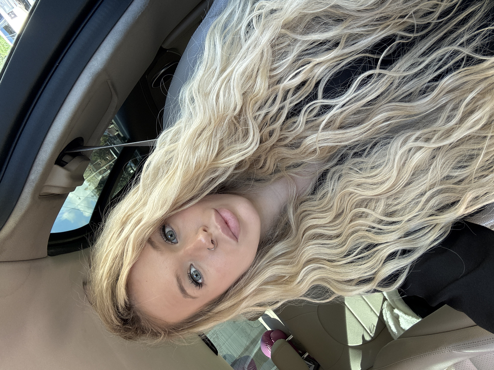 The media file is a portrait of a young woman with long, wavy blonde hair, taken inside a car. The camera is positioned to capture her face at an upward angle, with her hair cascading over her shoulder and across the frame. She is looking directly at the camera, with a neutral expression. Her eyes are blue, and she has a nose ring. The interior of the car is visible, with beige leather seats and a dark headliner. A seatbelt buckle is visible on the left side of the frame. Sunlight streams in from an open sunroof, casting shadows on the interior. The background outside the car is blurred, but appears to be a sunny day with some greenery and blue sky.
The image appears to be a selfie taken by the subject. The lighting suggests it is daytime. The scene is consistent with being in a vehicle. No visible text is present in the image.