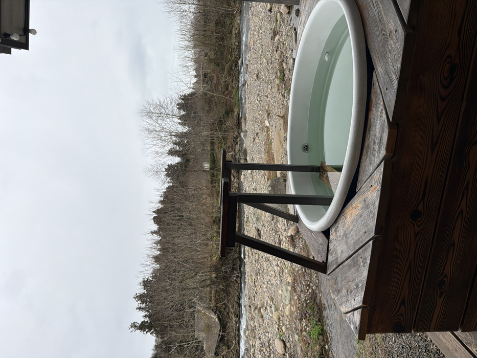 The image depicts an outdoor scene in Tsinsvla, Georgia. In the foreground, a white, oval-shaped bathtub, partially filled with light green-tinted water, is set into a dark, weathered wooden deck. The tub includes a visible drain and an internal wooden step. A dark, rustic wooden support structure, featuring two angled posts and a horizontal crossbar, stands over the tub. The wooden deck planks display prominent grain and show signs of weathering. Beyond the deck, a rocky riverbank slopes down to a river with moderate flow, visible ripples, and numerous exposed rocks. A smaller stream or inlet is present on the left side of the riverbank. The background consists of a dense line of trees, primarily bare deciduous trees, interspersed with some darker coniferous trees, under a uniformly overcast, light gray sky. No individuals or explicit actions are depicted within the scene.