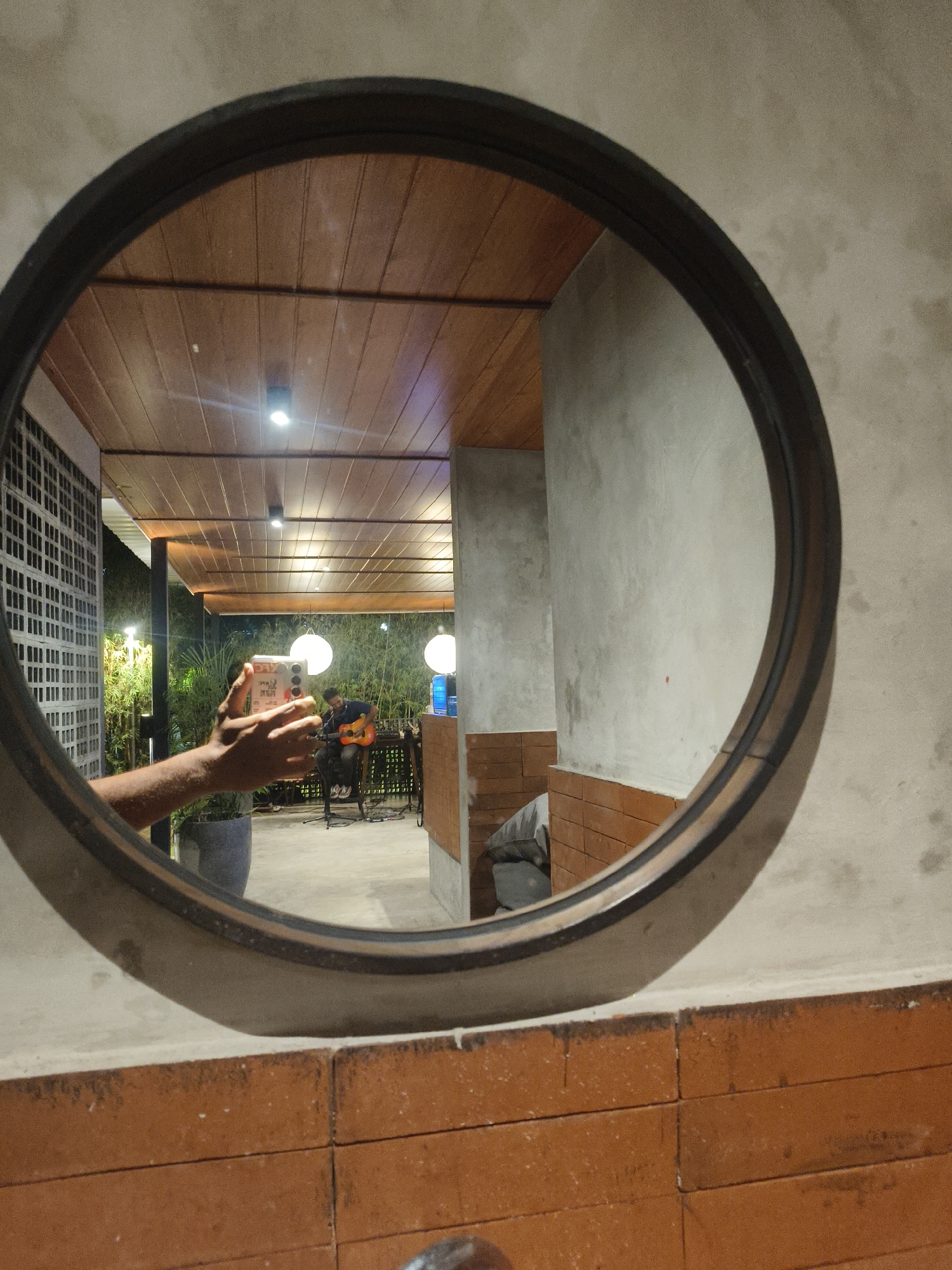 A circular mirror reflects a scene in Kuta Selatan, Indonesia. In the foreground of the reflection, a person's left arm and hand hold a white smartphone, angled to capture the reflection. The person appears to be standing near a large potted plant. Behind them, a man is seated, playing an acoustic guitar, with microphones and audio equipment visible. The background shows an outdoor or semi-outdoor setting with a wooden slatted ceiling, modern lighting fixtures, and lush green bamboo plants. The mirror itself is framed by a dark circular rim and mounted on a textured wall, with a section of orange brickwork visible below it.