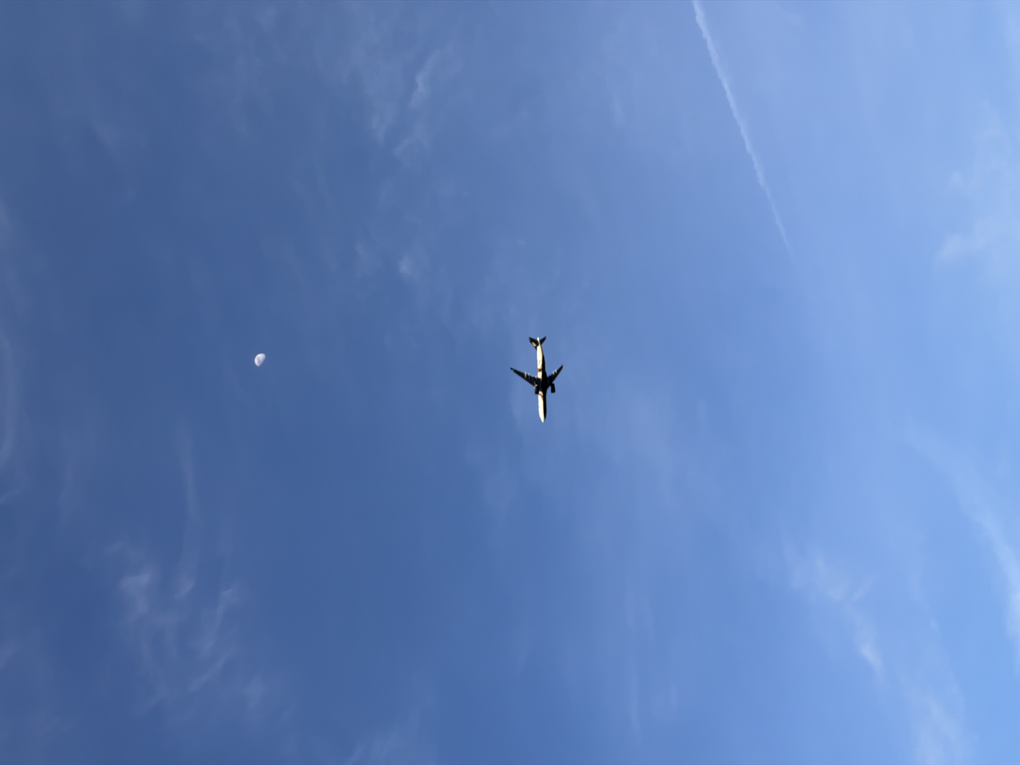 The image depicts an airplane flying in a clear blue sky. There is a faint contrail visible in the upper right portion of the image. A crescent-shaped object, possibly the moon, can be seen in the upper left. The photograph was likely taken in an outdoor setting. Without further information, it's not possible to determine if this was taken in Montreal, Canada.