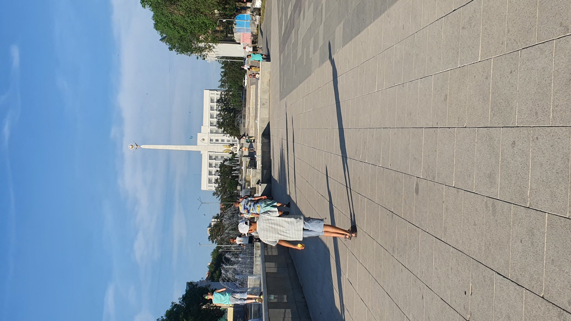 The image shows a sunny day with a clear blue sky in a city square. In the foreground, a person in a striped shirt and shorts stands with their back to the camera, holding a small yellow object. Behind them, another person in a brightly colored patterned shirt and shorts is visible. Further in the background, several other people are seen walking or sitting on a tiered structure. A tall, white monument with a star-like ornament at the top stands prominently in the middle ground, with a modern white building behind it. Trees and a fountain are also visible. The ground is paved with large grey tiles, casting long shadows from the sun, indicating it is likely late afternoon. The overall atmosphere appears relaxed and casual, suggesting a public space being enjoyed by visitors on a pleasant day. The location is identified as the Qarağandı city administration in Kazakhstan.