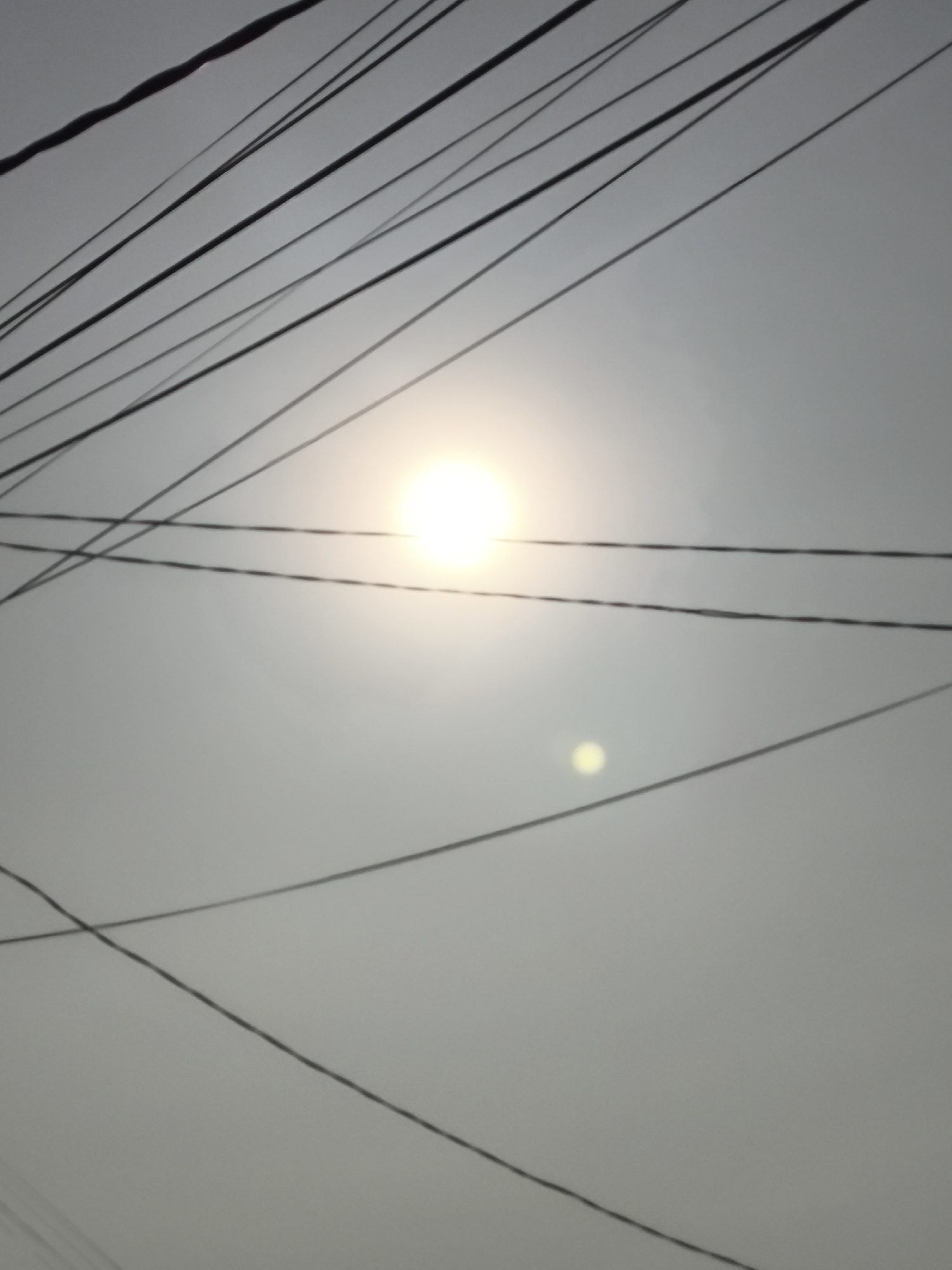 A low-angle, hazy photograph shows multiple power lines crisscrossing a muted, overcast sky. A bright, diffused sun is visible in the upper center of the frame, casting a soft glow. A small, circular light artifact is present in the lower right quadrant, likely a lens flare or reflection. The power lines are thin and black, creating a complex, layered pattern against the pale sky. The image appears to have been taken outdoors in an urban or suburban environment with abundant overhead wiring. The location in Cilegon, Indonesia, is suggested by the presence of such dense power line infrastructure, common in many parts of the country.