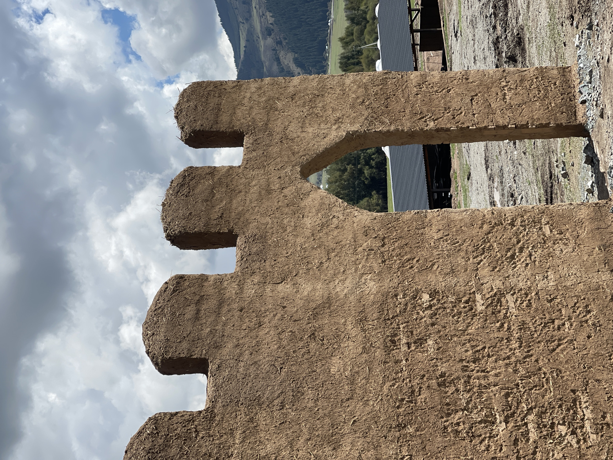 In Semenovka, Kyrgyzstan, a partially constructed adobe wall with crenellations at the top stands prominently. The wall features an arched opening, revealing a rural landscape beyond. In the background, agricultural fields and forested hills are visible under a cloudy sky. Several dark-roofed structures, possibly stables or storage buildings, are situated near the wall. The ground in the foreground is uneven and appears to be a mix of dirt and gravel. The overall scene depicts an unfinished construction project in a natural, mountainous environment.