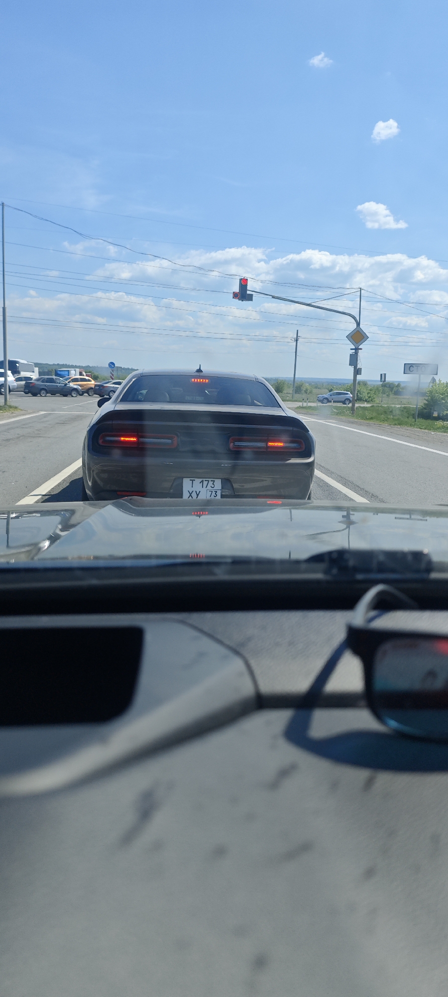 A rear view of a dark-colored Dodge Challenger with the license plate "T 173 XY 73" is visible through the windshield of another car. The Challenger is stopped at a traffic light in Prigorodny, Russia. The traffic light is red. The sky is mostly clear with scattered clouds. Other vehicles are visible in the background. Road signs are present on the right side of the road. Sunglasses are on the dashboard of the car taking the photo.