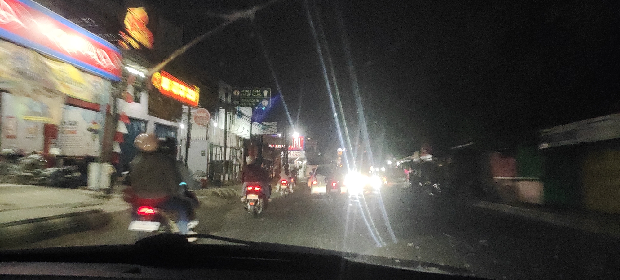 The image depicts a night scene on a street in Tanubayan, Indonesia, likely a commercial area given the presence of illuminated signs and shops. The perspective is from inside a vehicle, as indicated by the windshield frame in the foreground. Several motorcycles are visible, with riders on them, suggesting traffic or movement on the street. One motorcycle in the foreground has its brake light illuminated. Buildings line the street, some with bright, neon signs, one of which appears to be red and yellow. A street sign pointing to "DEMAK KOTA MASJID AGUNG" and "SRWODADI" is also visible. The overall lighting is dim, with artificial lights from signs and vehicle headlights creating a strong contrast. Some areas of the image are overexposed due to bright lights, and there are light streaks, possibly from streetlights or headlights, across the scene. The environment appears to be a city street at night.