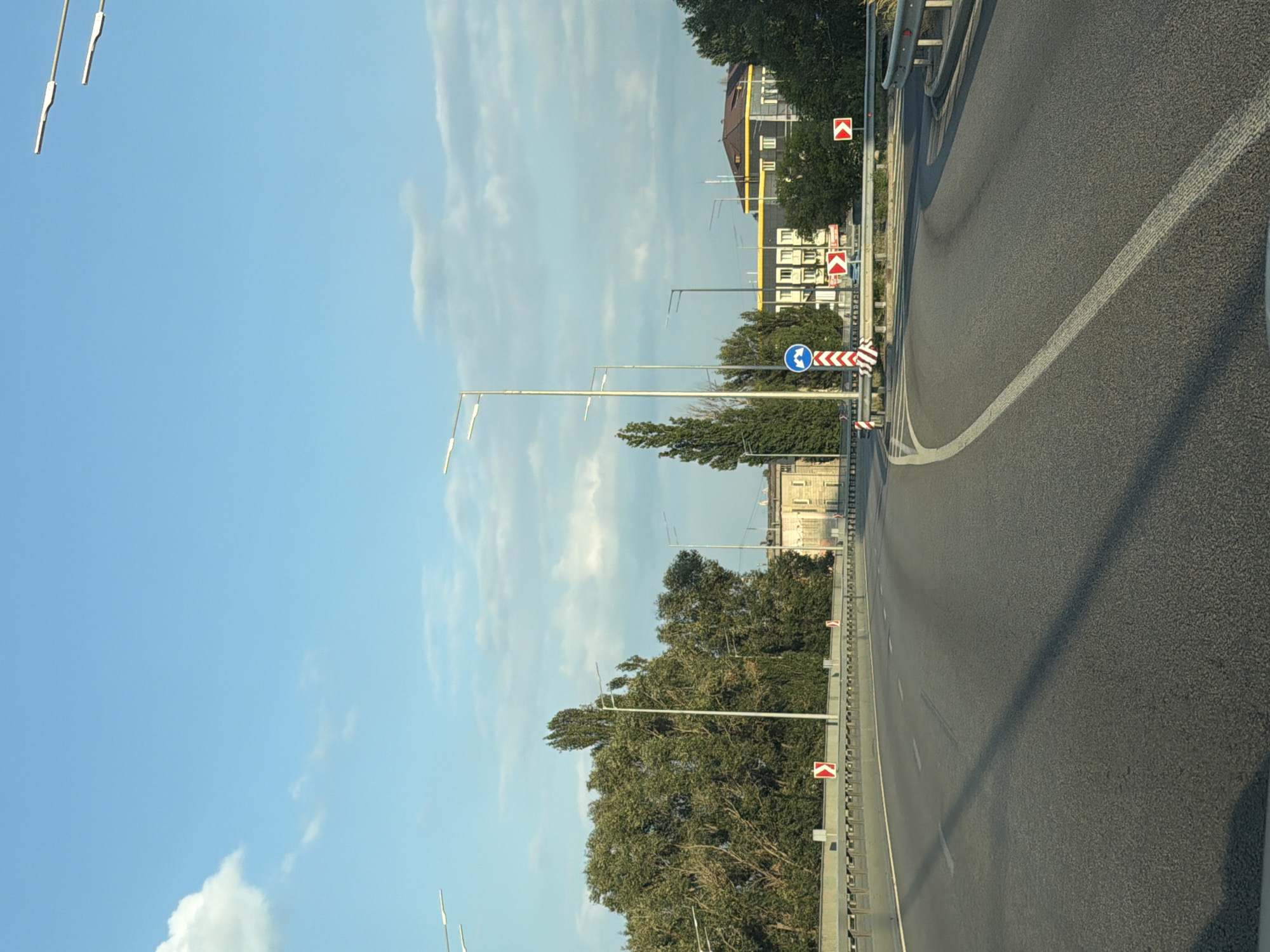 The image depicts a highway scene in Kyiv, Ukraine, on a clear day with a blue sky dotted with white clouds. The road, a multi-lane asphalt surface, curves to the right, with white lane markings. Guardrails line the sides of the road, and several streetlights are visible, stretching across the frame.
On the left side of the road, a dense cluster of trees with lush green foliage is present. Further in the distance, a building with a beige facade and a lighter colored roof is visible.
On the right side of the road, several traffic signs are placed. These include a directional sign indicating a curve to the right with a chevron symbol, and a circular blue sign with two white arrows, suggesting a recommended route or direction. There are also more red and white chevron signs along the guardrail, indicating a road hazard or turn. In the background on the right, a modern building with a dark roof and light-colored walls is observable, surrounded by trees. The overall environment suggests a well-maintained roadway within an urban or suburban area.