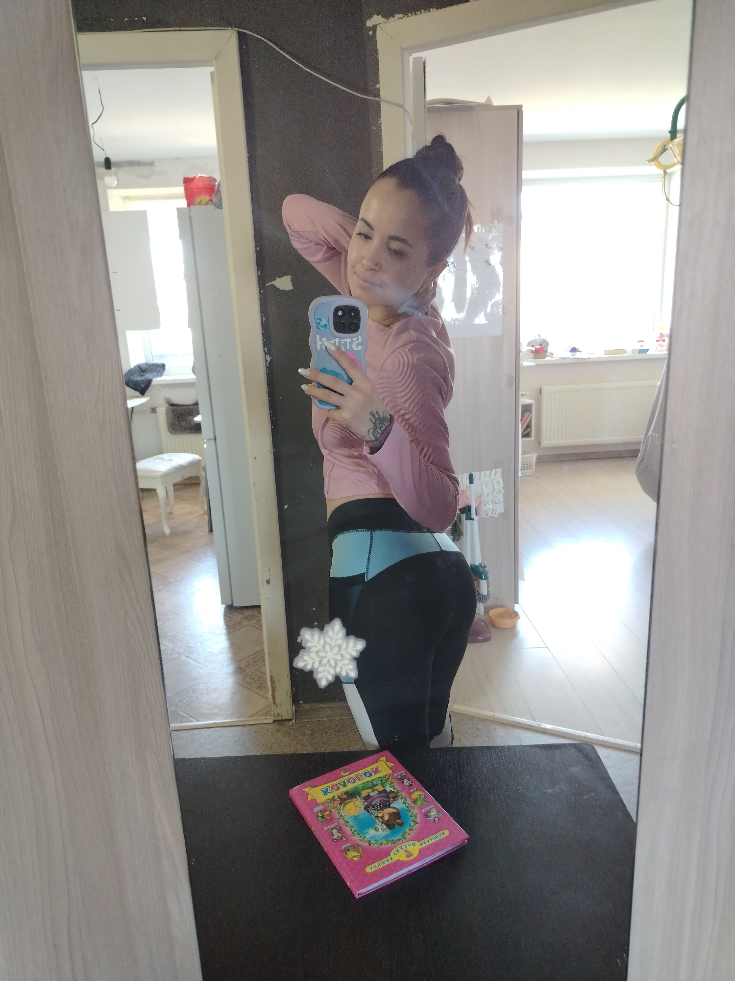 A young woman takes a mirror selfie in what appears to be an apartment. She is wearing a pink long-sleeved shirt and black leggings with blue accents. Her hair is pulled back in a bun. She is holding a blue smartphone. The mirror reflects two rooms: one with a white stool and the other with a radiator. There is a pink book on the surface in front of the mirror. There is a snowflake decoration attached to the mirror.
It is impossible to confirm the city is Noviy, Russia based on the image alone.