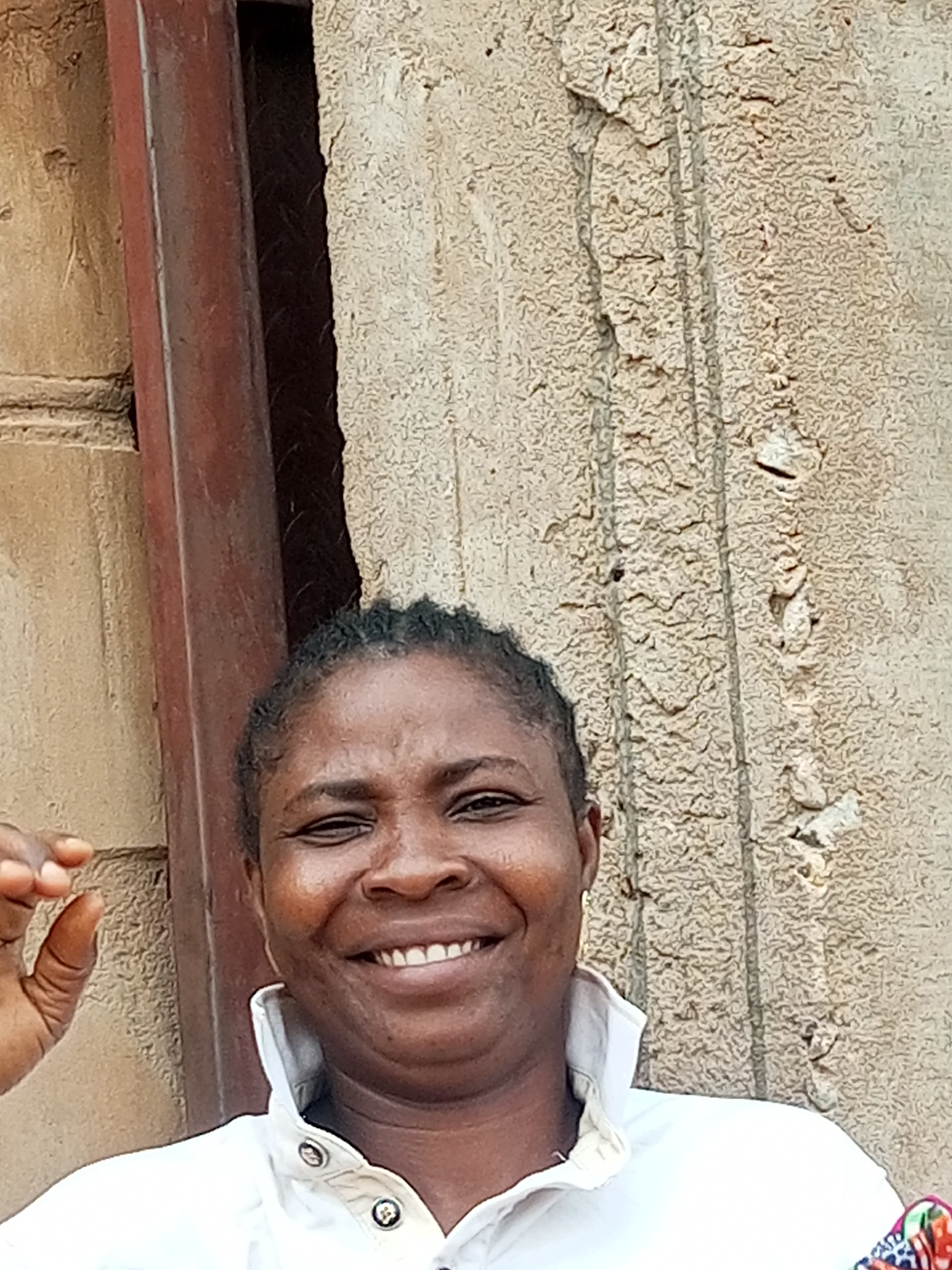 A medium shot photograph captures a dark-skinned woman with her hair in braids, smiling broadly. She is wearing a white collared jacket adorned with decorative buttons. Her left hand is raised, with her index finger and thumb forming a small circle. The background consists of a textured, beige-colored wall and a portion of a dark, reddish-brown vertical support beam. The scene appears to be outdoors, possibly on a street or in a courtyard within Tempure, Nigeria.