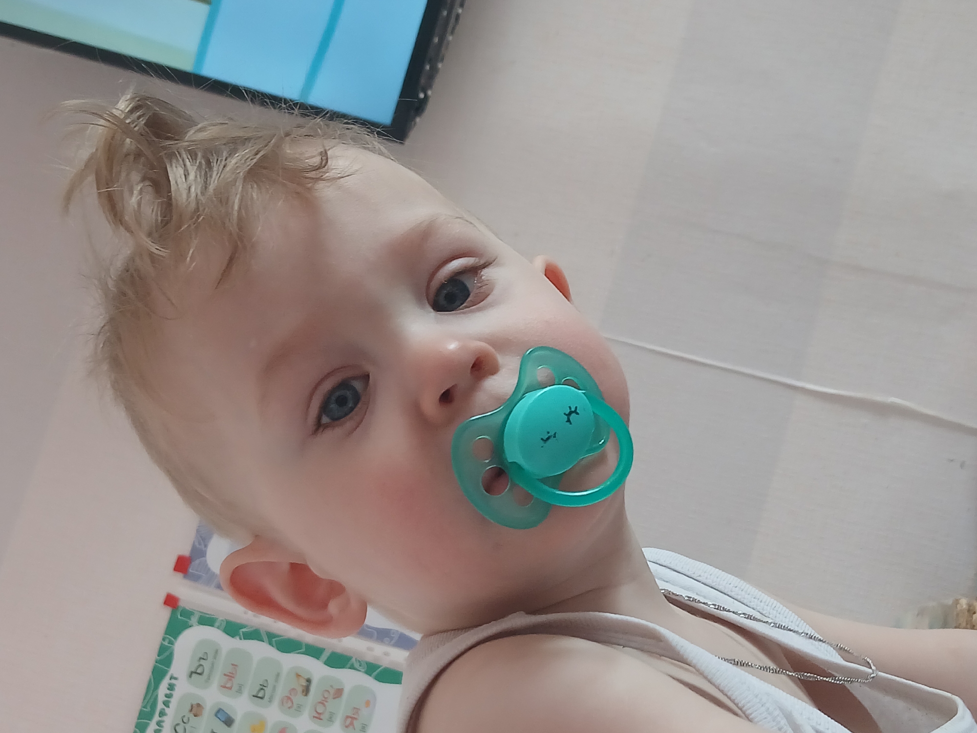 A close-up, slightly angled shot shows a light-skinned baby with blue eyes and blonde hair. The baby is facing the camera with a green pacifier in their mouth. They are wearing a white tank top, and a thin silver chain necklace is visible around their neck. In the background, blurred, is a blue television screen in the upper left and what appears to be a white wall or surface. A colorful board or book with what appear to be Cyrillic letters on it is visible in the lower left corner. The image suggests an indoor setting, potentially a home. There is no discernible information in the image to establish the location of the image within the city of Chaikovsky, Russia.