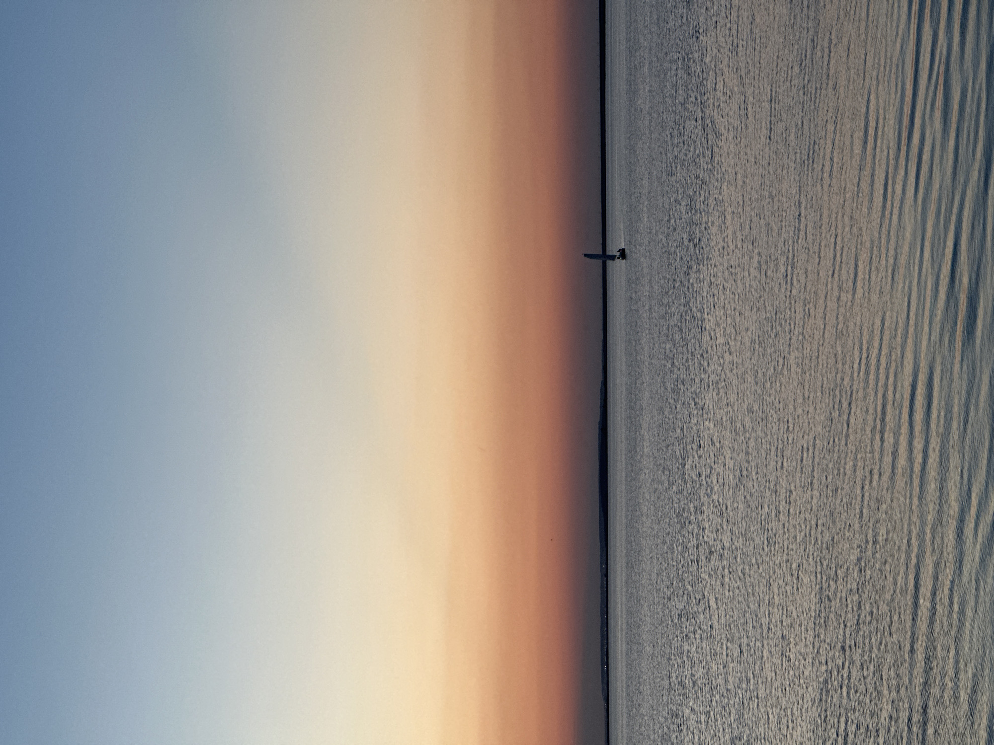 The image depicts a serene sunset over a body of water, likely a lake, in Kokshetau, Kazakhstan. The sky transitions from a pale blue at the top to soft hues of orange and pink closer to the horizon. A faint landmass is visible in the distance, silhouetted against the colorful sky. On the water, a small sailboat with a single sail is visible, moving gently across the surface. The water itself is calm, with subtle ripples reflecting the colors of the sky. The overall atmosphere is peaceful and tranquil.