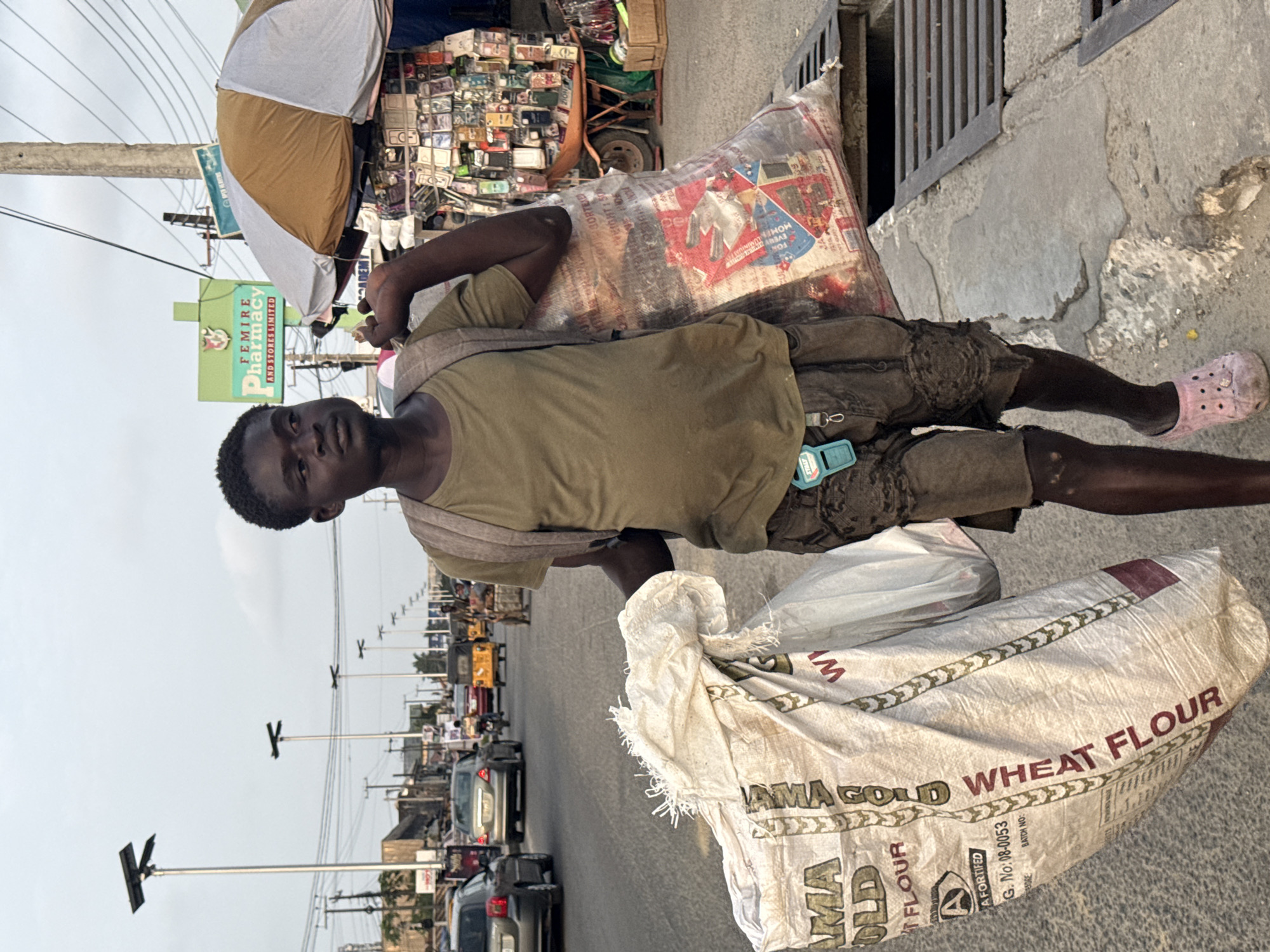 A young Black male walks along a city street in Ado, Nigeria, carrying two large bags. He wears a light olive-green t-shirt and dark, distressed shorts. A light brown backpack is slung over his shoulders. In his right hand, he holds a light-colored, woven bag with red and gold lettering that reads "AMA GOLD WHEAT FLOUR". To his left, he carries a translucent plastic bag filled with various items, some appearing to be food products. He wears a pair of light pink clogs on his feet. The street appears busy with traffic, including cars, a yellow auto-rickshaw, and motorcycles. Various businesses, including a "FEMIRE PHARMACY AND STORES LIMITED", line the street. Overhead, streetlights and power lines are visible. The ground is paved with concrete, and a metal grate, likely a storm drain, is present near the man's feet. The overall lighting suggests it is either early morning or late afternoon.