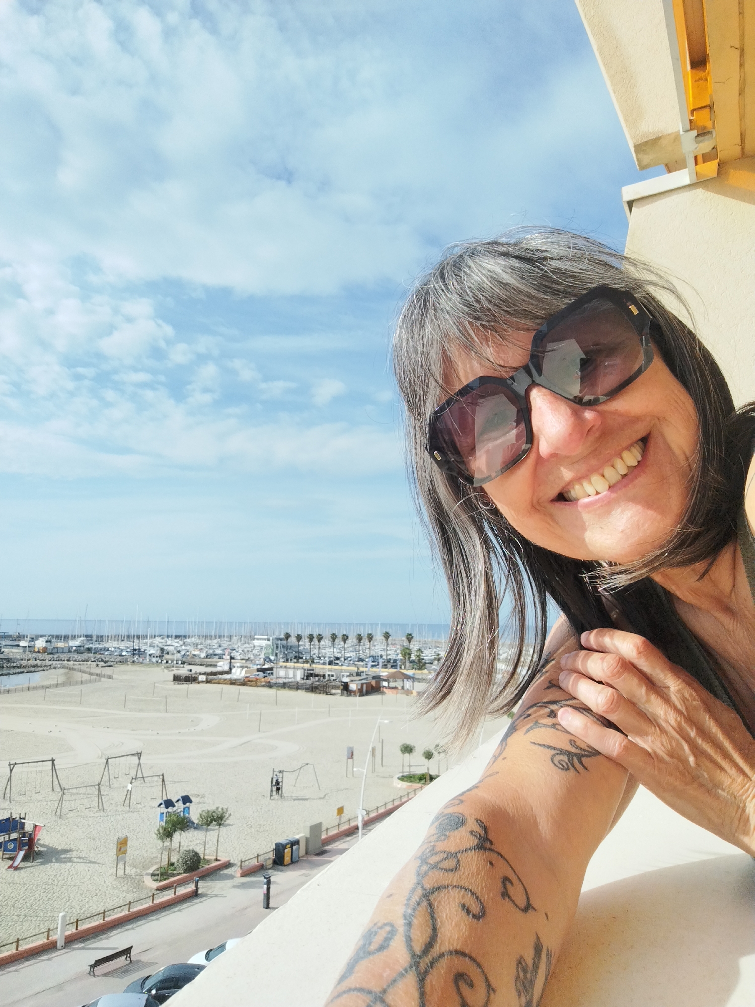 A woman wearing sunglasses is smiling into the camera on a sunny day. The location appears to be Palavas-les-Flots, France, based on the harbor, beach, and cityscape visible in the background. The beach is sandy with playground equipment and a few people present. A marina filled with boats and yachts is visible in the distance. Palm trees line the harbor. The woman has gray hair and a tattoo on her arm. She is standing on a balcony, evidenced by the railing.
