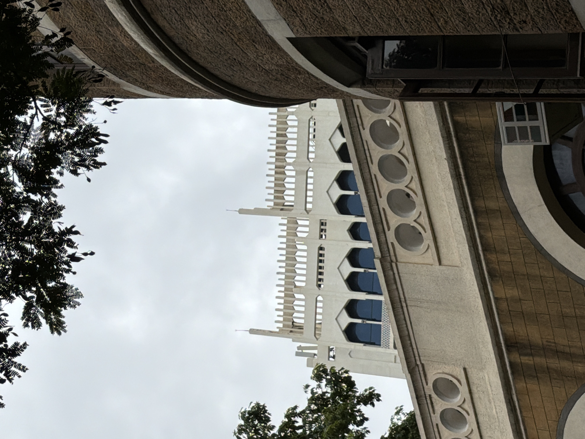 This is a low-angle shot of the exterior of a building, possibly an old colonial-era structure, in Mumbai, India. The architecture is a mix of styles, with a prominent light-colored, ornate upper section featuring pointed arch windows and decorative crenellations, contrasting with a darker, more solid stone lower section. A curved, brown brick tower-like element dominates the right side of the frame, showing a section of an open wooden window. Overhanging tree branches with dark leaves partially obscure the sky, which is overcast and a pale gray. The scene suggests an institutional or historical building, likely in a well-established part of the city. There are no people visible.