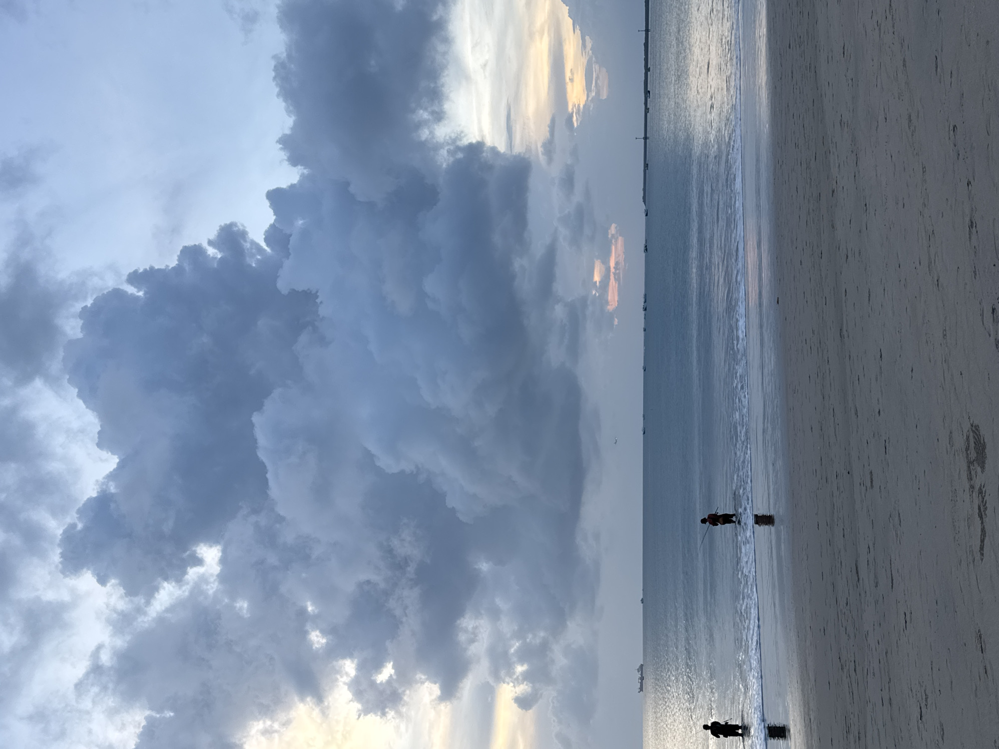 The image shows a beach scene in Kuta, Indonesia. The sky is dominated by a large, dark, cumulonimbus cloud formation, tinged with some sunset colors, near the horizon. Below the cloud, the ocean meets the sandy beach. Two people are standing near the water's edge. The scene appears to be at dusk or dawn.