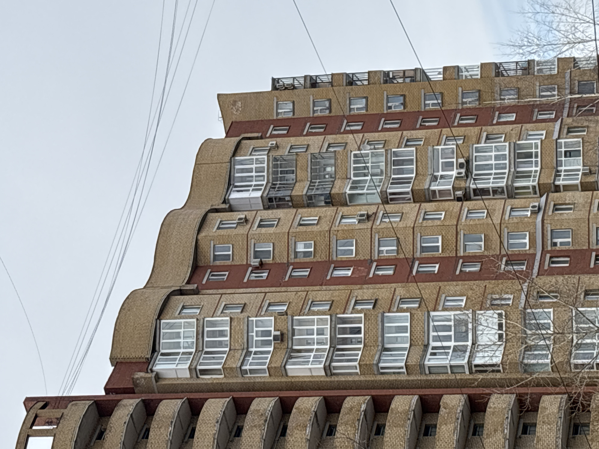 The image shows a close-up of a distinctive building in Perm, Russia. The architectural style is characterized by its tiered, terraced design with a consistent repetition of window units. The building's exterior is primarily beige brick, with dark red horizontal accents between levels. The windows are framed in white, with large, protruding glass sections that extend outward. On the upper levels, there are smaller, rectangular windows. The top of the building features a terrace-like area with railings. In the background, thin wires are visible against a pale sky. The bottom right of the frame includes a few bare tree branches.
