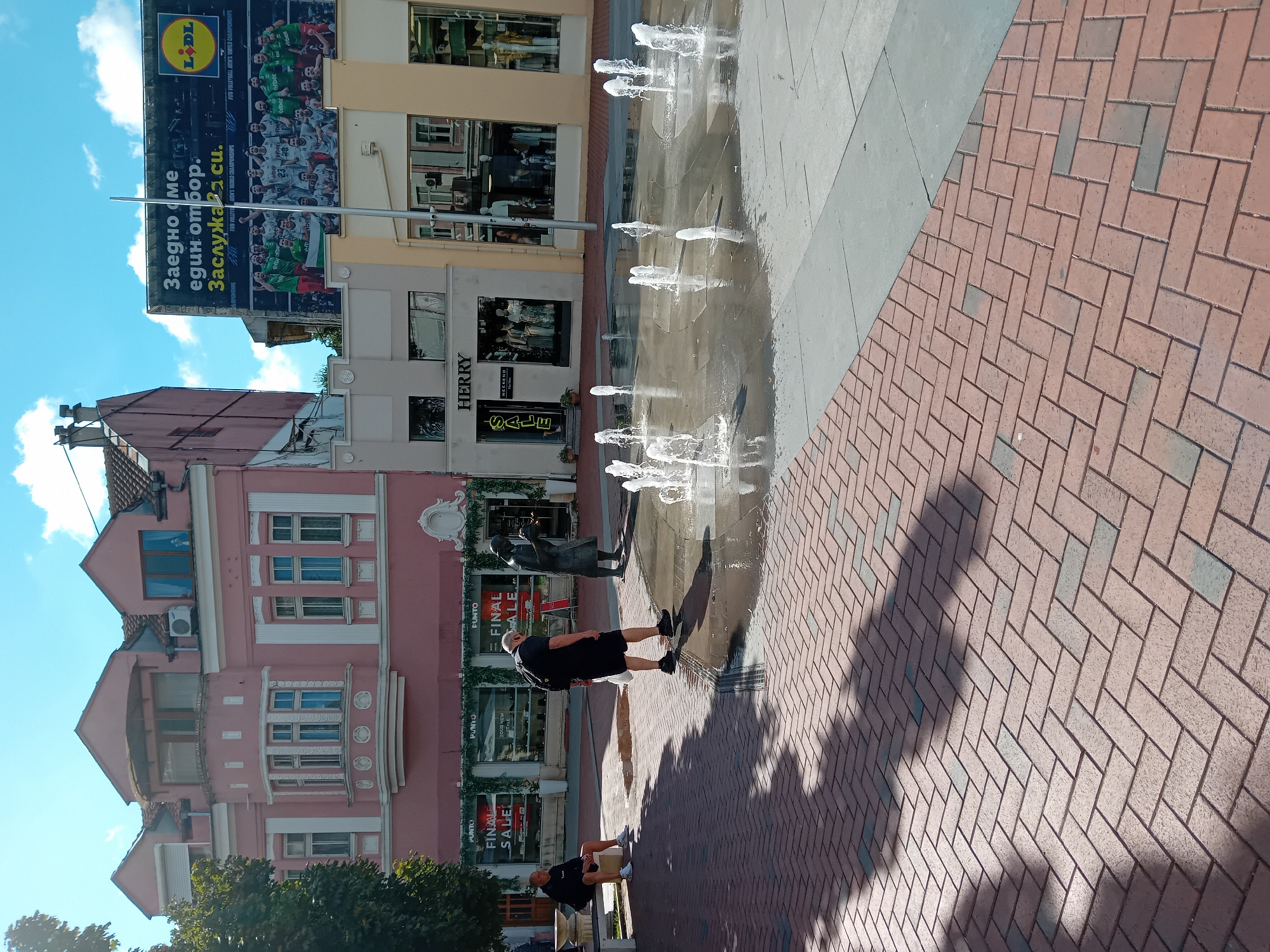 In Varna, Bulgaria, on a sunny day, a paved pedestrian area features a shallow water fountain with multiple jets. Two men are present; one sits on a white bench to the left, facing away from the camera, wearing a black shirt and white shorts. The second man, in a black t-shirt and shorts, walks towards the fountain from the left, carrying a white plastic bag. He is casting a shadow onto the red brick herringbone-patterned pavement in the foreground.
Behind the fountain, a bronze statue of a woman reading a book stands. Further back, storefronts line the street. To the left, a pink building with white trim has multiple windows and balconies. One storefront displays "PUNTO" signage and "FINAL SALE" banners. Adjacent to it, a grey building has "HERRY" lettering above its windows. To the right, a yellow-accented building shows mannequins in its display windows.
Above the buildings, a large advertisement billboard displays a group of athletes in green and white uniforms, with text in Bulgarian and the LIDL logo. The sky is blue with scattered white clouds. The environment suggests a commercial or recreational urban space.