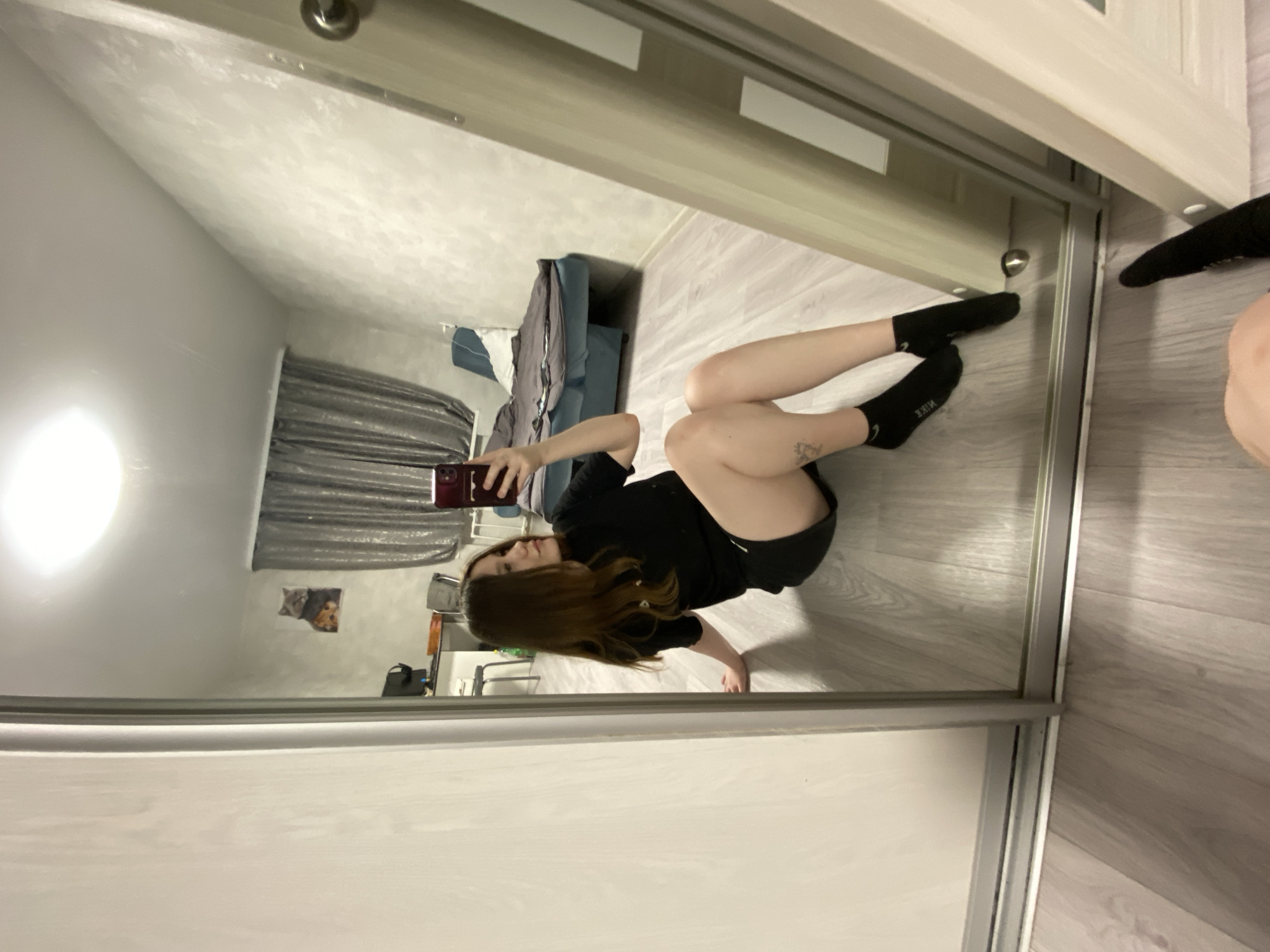 This image is a mirror selfie taken by a woman lying on the floor in an indoor setting. The photograph is oriented 90 degrees clockwise, making it appear as if the woman is on a wall, but she is reflected lying on a light wood-patterned floor.
The main subject is a young woman with long, reddish-brown hair, wearing a black t-shirt, black shorts, and black socks with white text. She is holding a smartphone with a dark red case up to her face, looking at the screen as she takes the photo. A small, dark tattoo is visible on her left thigh.
The reflection reveals a room with white walls and a somewhat textured white ceiling. In the background, a dark, striped curtain covers what appears to be a window. A blue or grey upholstered furniture piece, possibly a bed or sofa, is also visible. On the wall behind the woman, a picture or poster featuring two cats is displayed. A bright, circular light source is reflected on the far left wall, suggesting artificial indoor lighting. The frame of the large sliding mirror, likely part of a wardrobe or closet, is visible, including a small metallic handle.
Notable details include the casual pose for the selfie and the clear view of the word "NIKE" written in white on her black socks. The location is Chaikovsky, Russia.